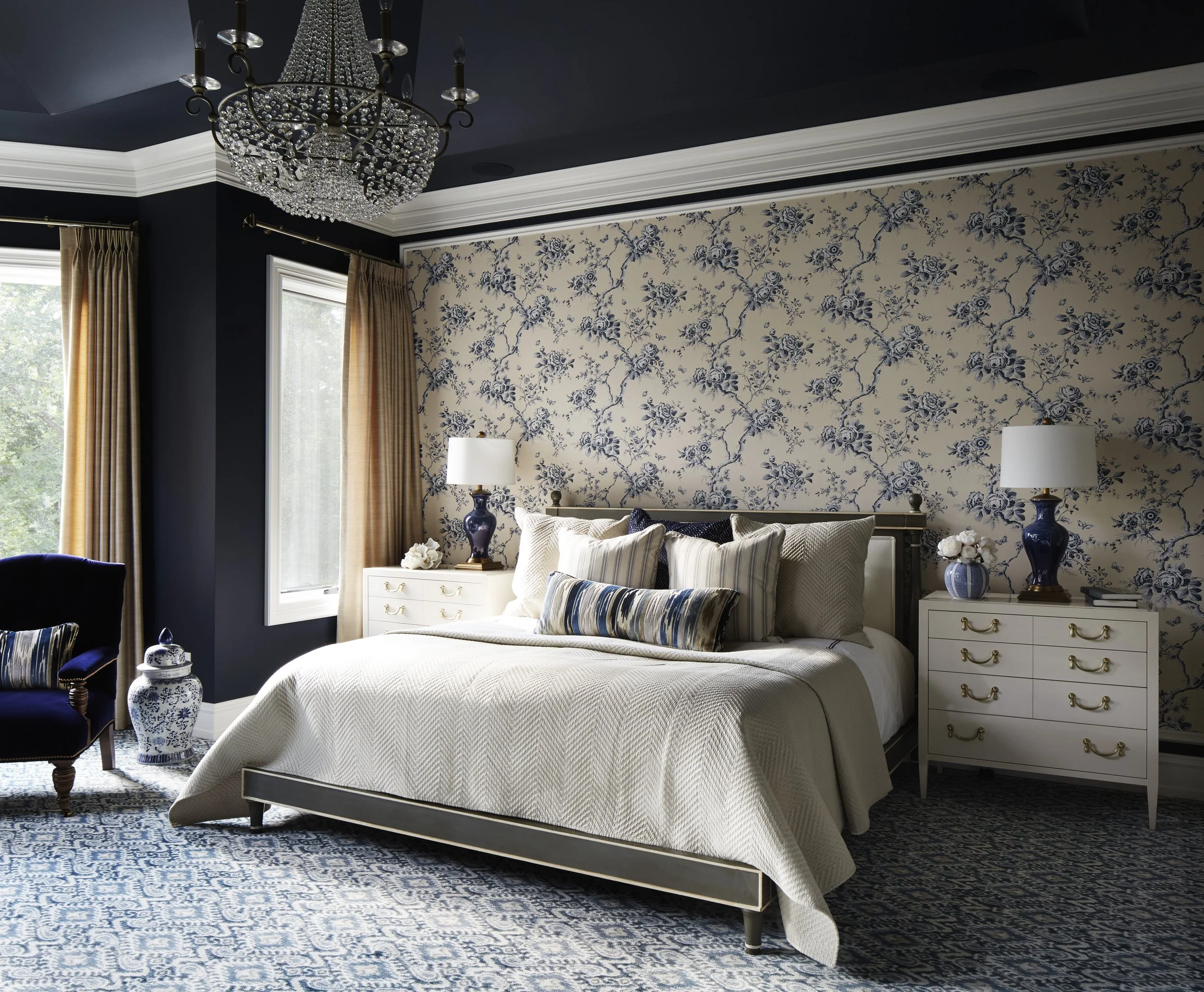 A bedroom with a blue and white floral wallpaper, a white bed with pillows, bedside lamps, a chandelier, and a blue patterned rug.