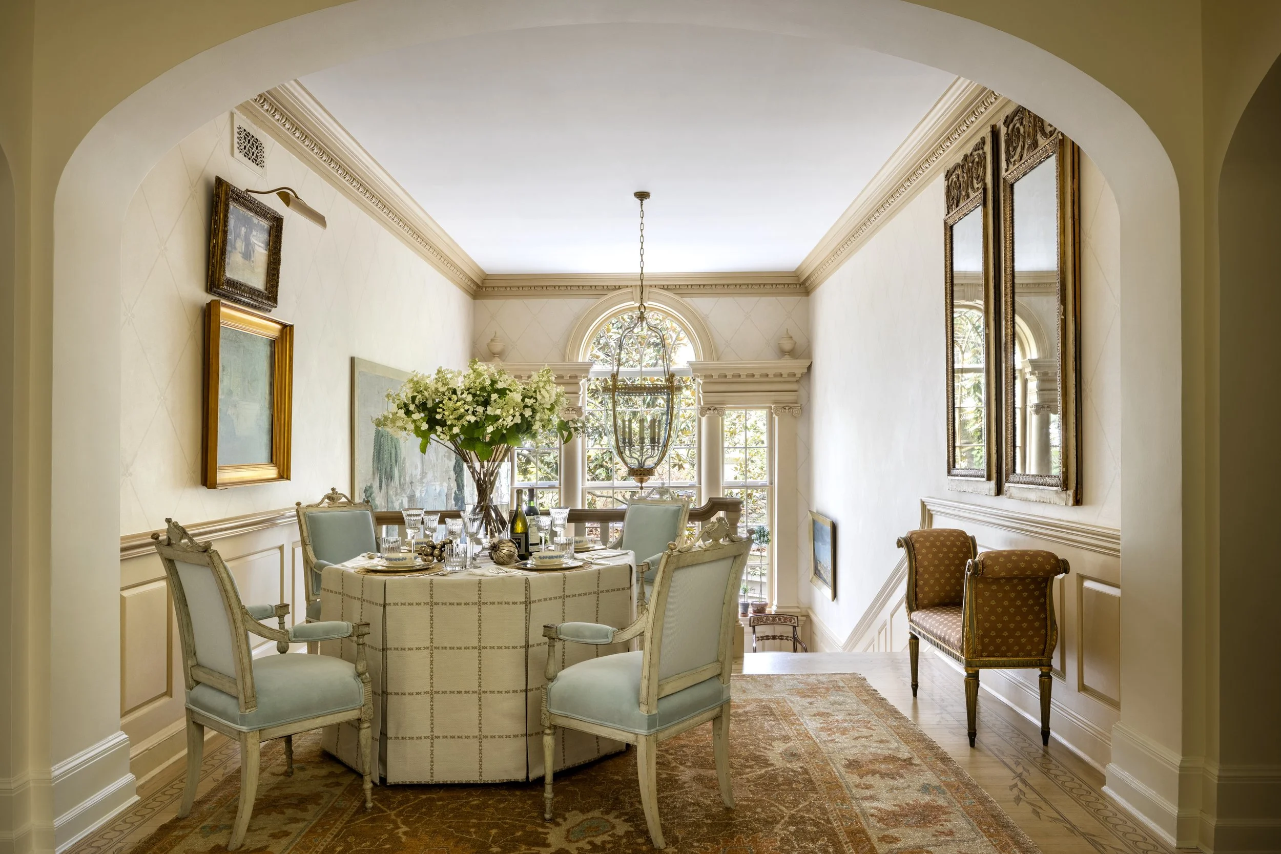 Elegant dining room with a round table set for a meal, surrounded by vintage armchairs, with floral centerpiece, overlooking large windows and decorated with framed art and ornate moldings.