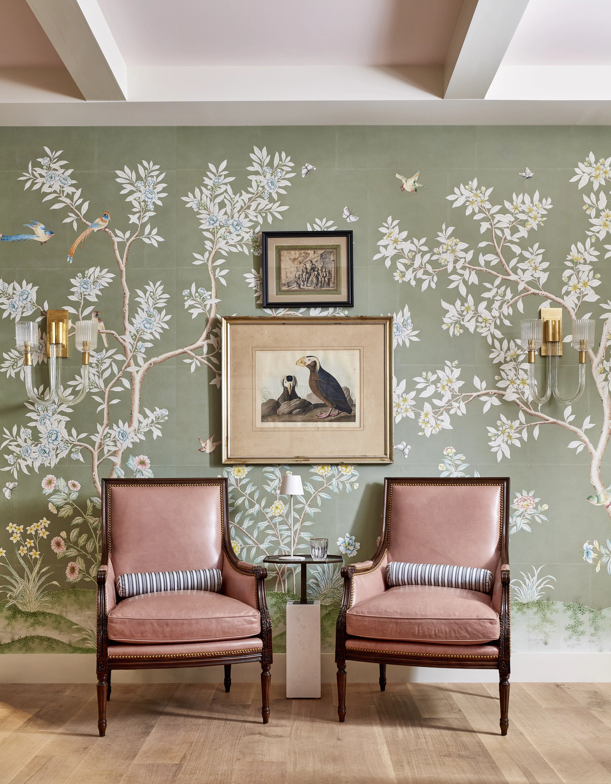 A room with two pink velvet armchairs with wooden frames and striped pillows, a small marble table with a glass and a lamp between the chairs, a green floral wallpaper with white flowers, hummingbirds, and butterflies, framed artwork on the wall depicting birds, and a hardwood floor.
