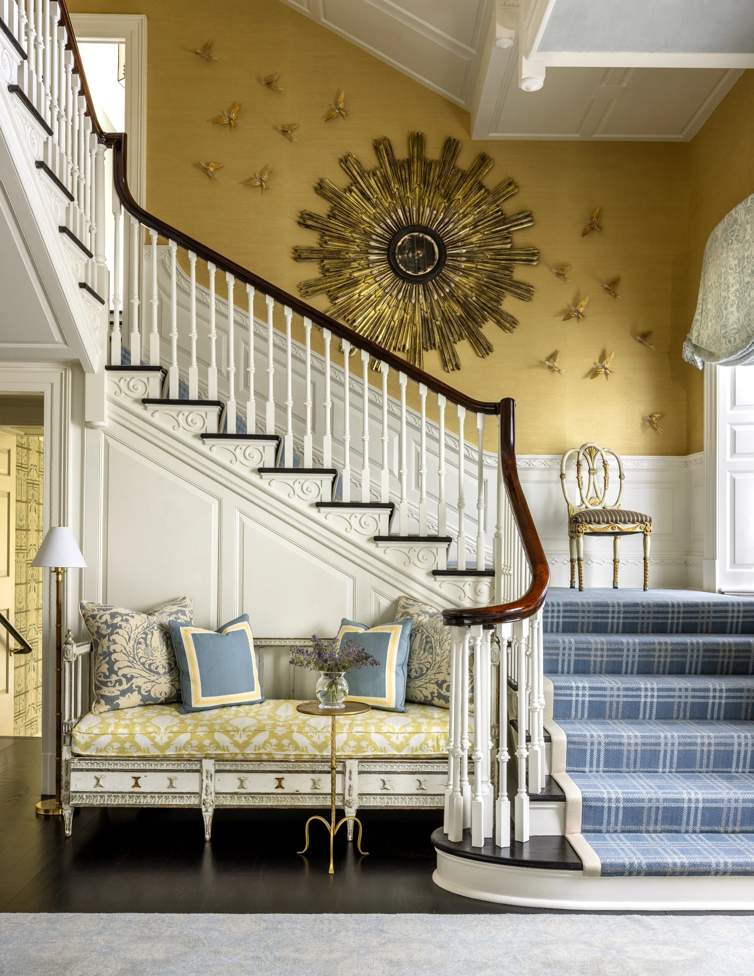 A staircase with blue and white striped carpeting, decorative white railings, and an ornate white and gold chair on the landing. Next to the stairs, there's a seating area with a yellow patterned sofa, plaid cushions, and a glass table with a flower 