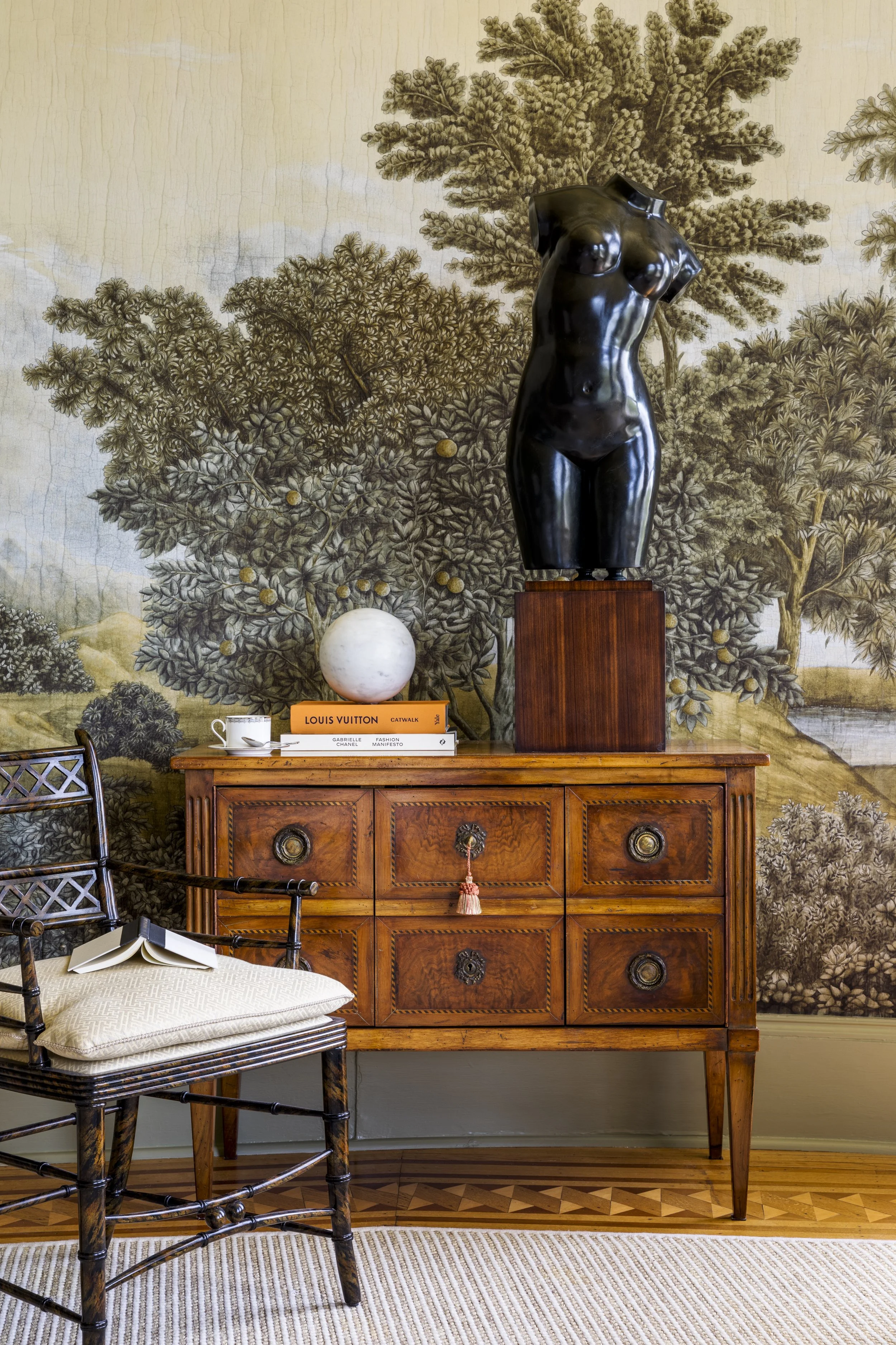 A black sculpture of a female torso on a wooden pedestal, set against a detailed botanical wallpaper. A wooden sideboard holds books, a white sphere, a teacup, and an open book. A black chair with a cream cushion and a striped rug are nearby.