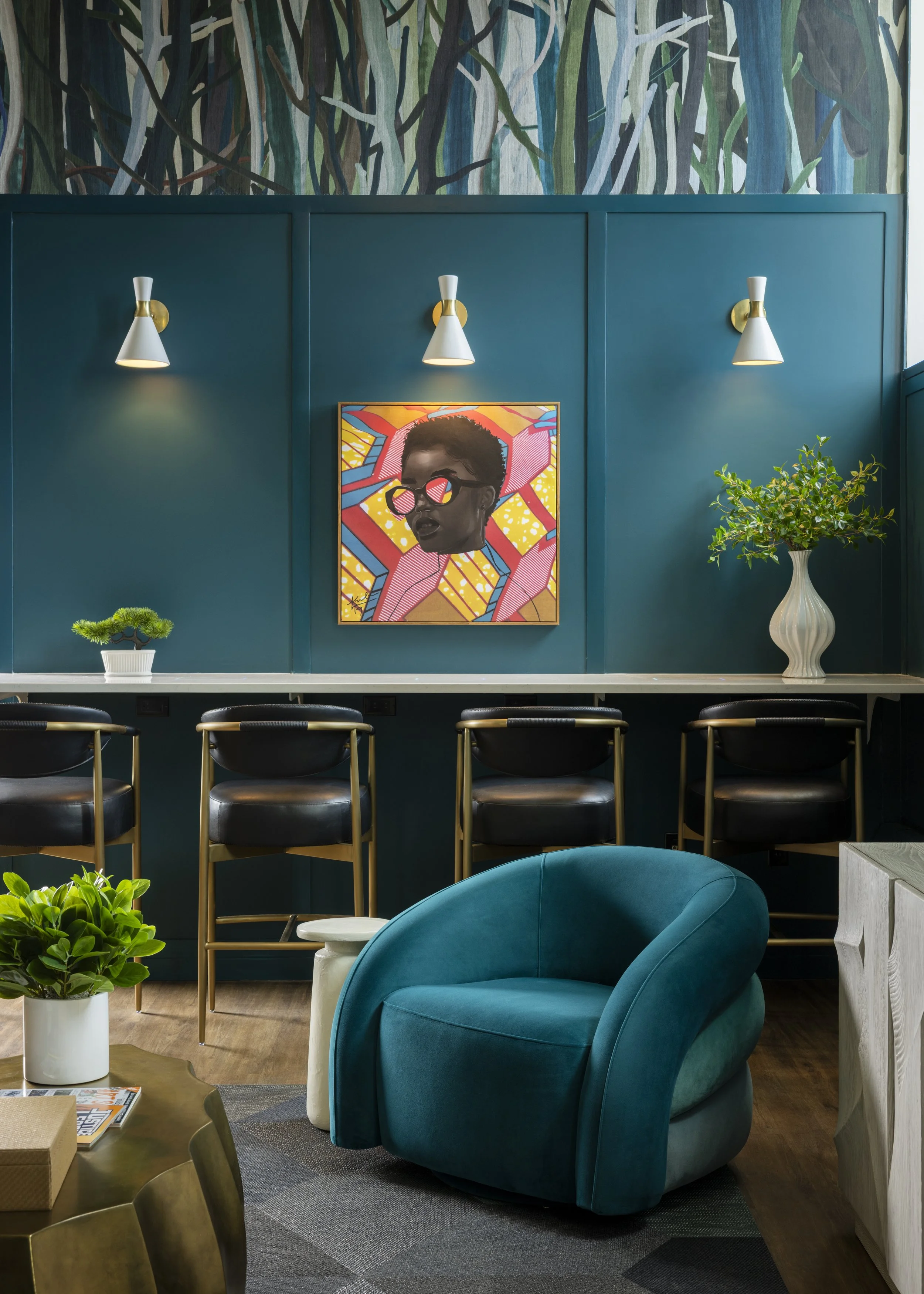 A stylish interior space featuring a teal wall with three wall lamps and a colorful pop art portrait of a woman wearing sunglasses. Decor includes a large white vase with a leafy green plant, a small bonsai tree, a teal velvet armchair, and a light-c