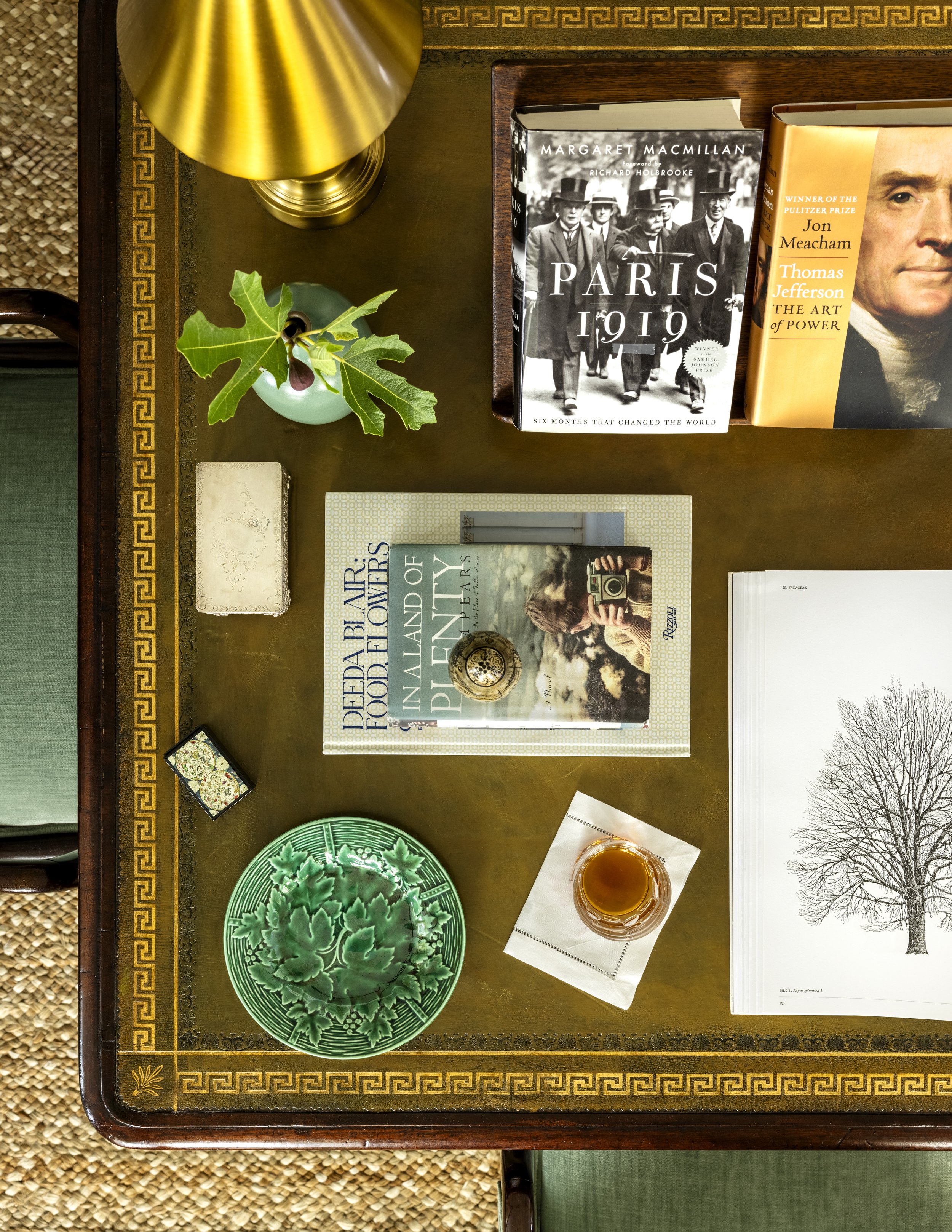 A wooden table with a decorative border, a gold table lamp, a small potted plant with green leaves, several books, a green decorative plate, a glass of amber-colored beverage, and a notebook with a tree illustration.