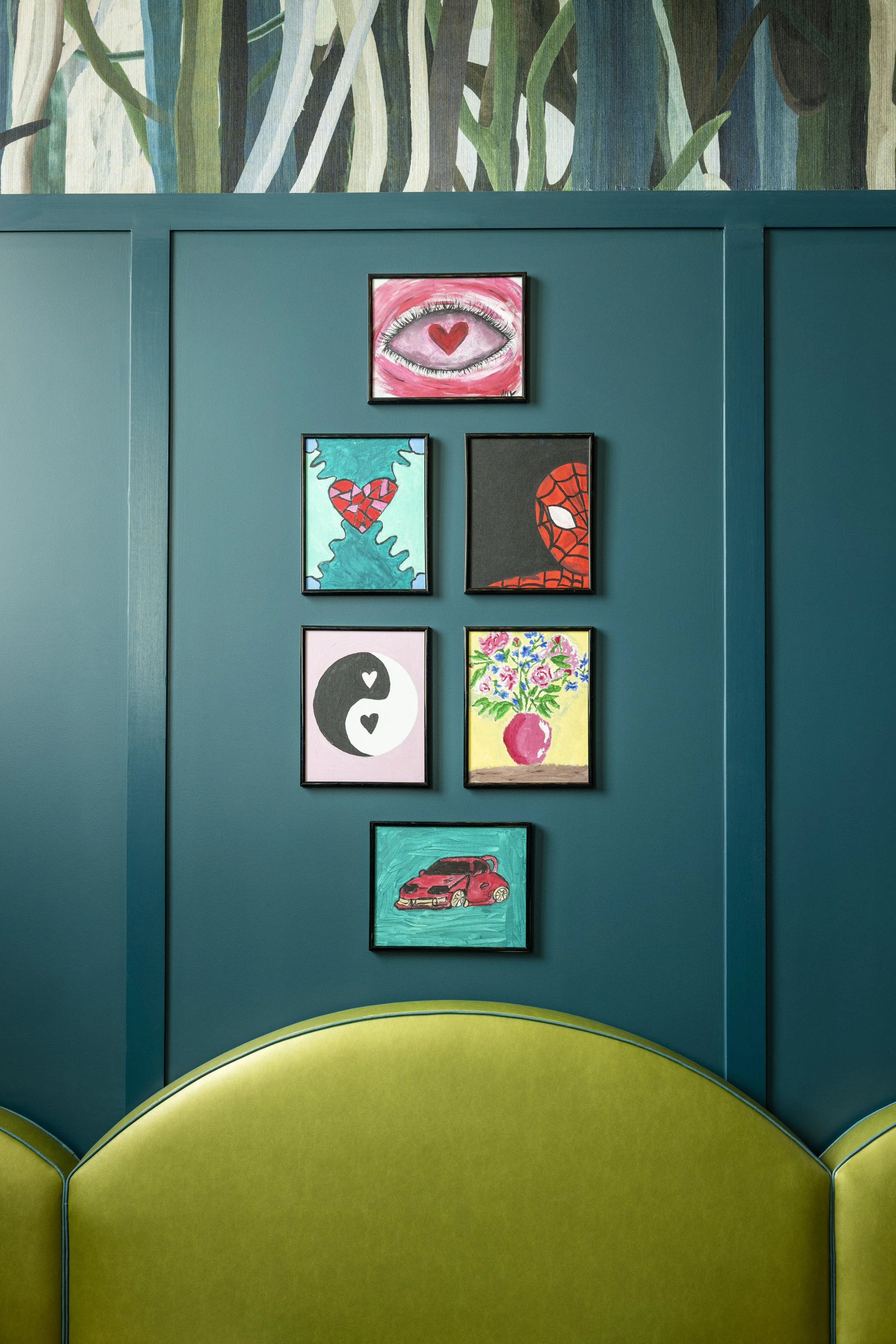 Green upholstered chair in front of a blue wall decorated with six framed pictures. The top picture depicts an eye with a red heart inside, the second row has a heart with blue and red colors and a face resembling Spider-Man, and the bottom row featu