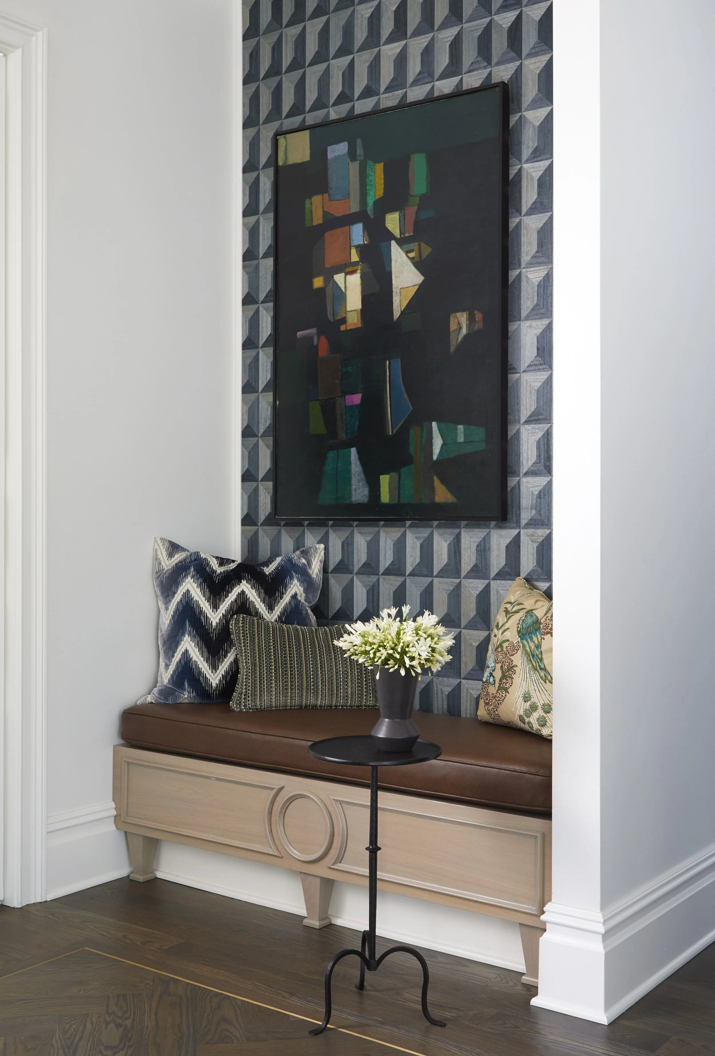 Living room seating area with a wooden bench underneath a geometric-patterned blue and gray wall, decorated with three cushions and a floral pillow, a vase with white flowers on a small round black table, and a large abstract painting on the wall.