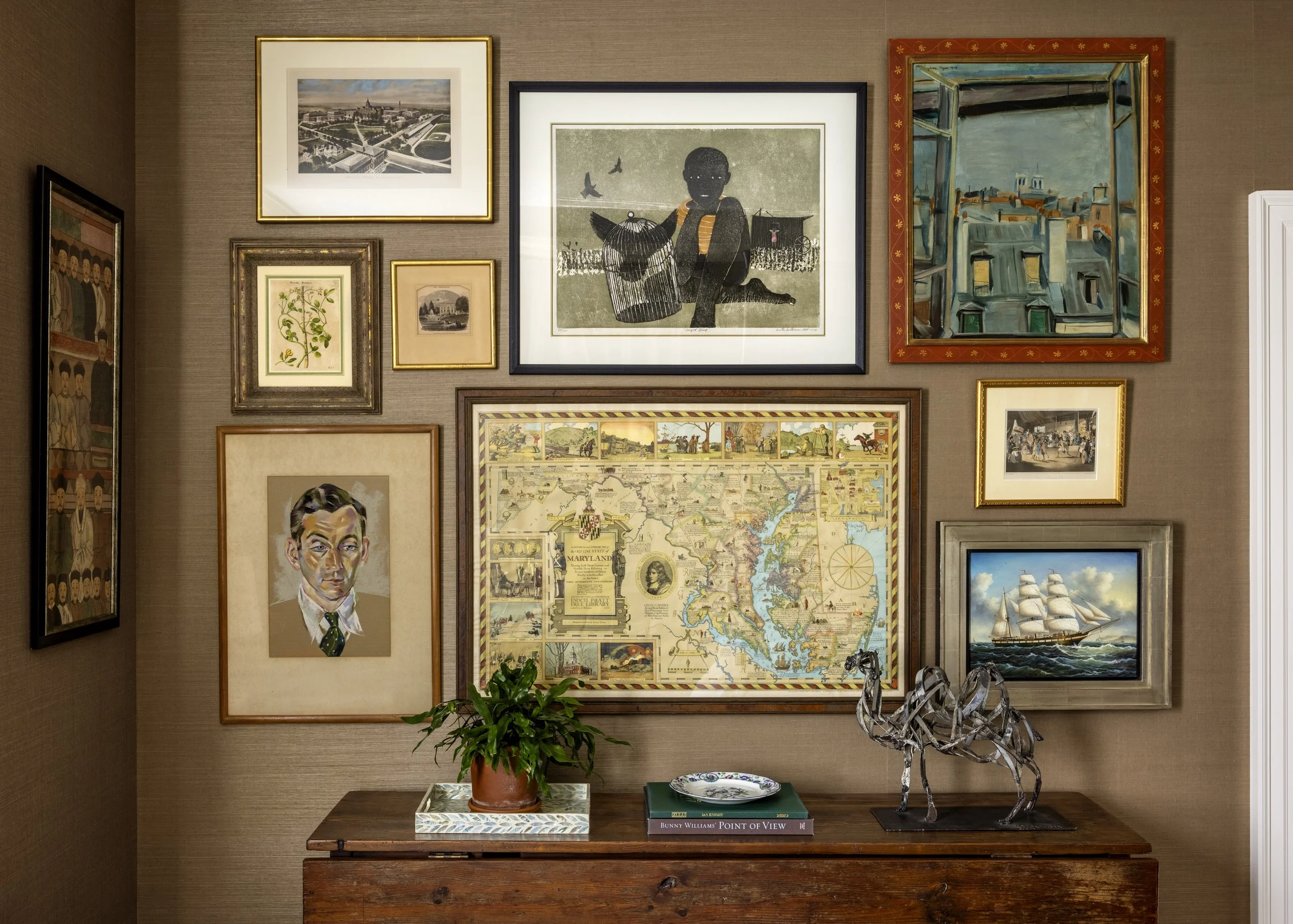 A gallery wall with various framed artworks including a cityscape, a child with birds, a map of Maryland, and other paintings. A wooden table below holds a potted plant, a decorative tray, and books. A metallic sculpture of a horse is on the right si