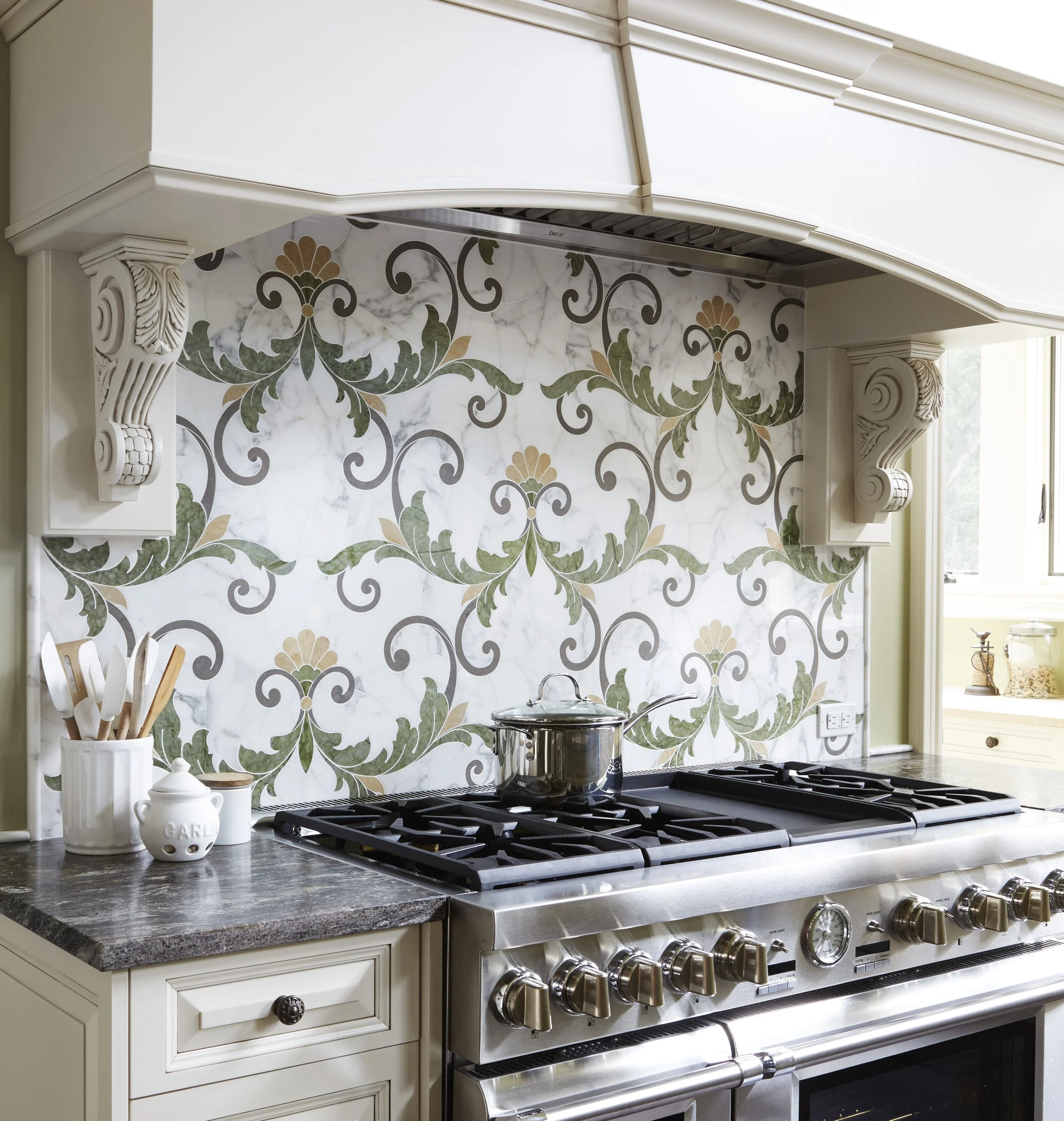 A kitchen stove with a stainless steel pot, decorative backsplash with floral patterns, white cabinets with ornate crown molding, and a container holding wooden cooking utensils.