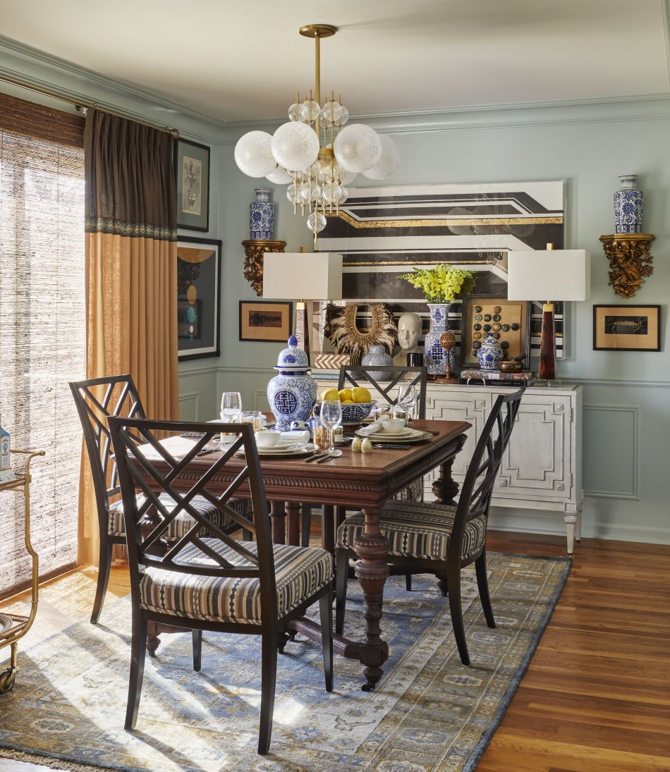 A decorated dining room with a wooden table, six chairs with striped cushions, a patterned area rug, and a buffet with vases, art pieces, and a lamp. A chandelier hangs from the ceiling, and there are curtains on the window.