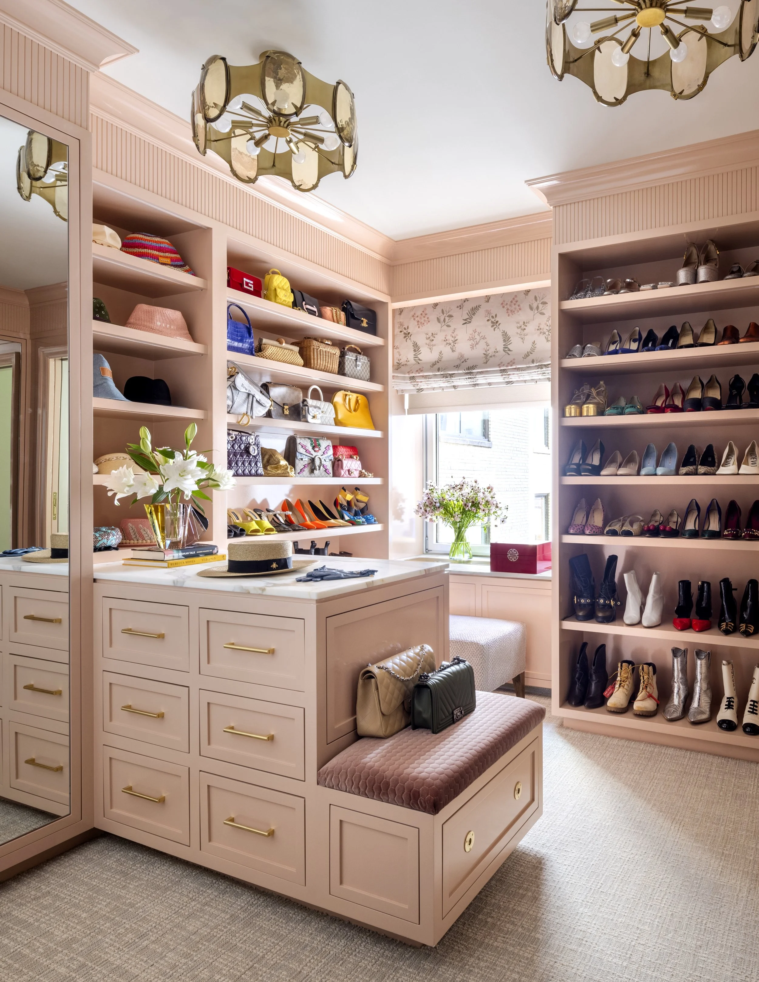 A walk-in closet with pink cabinetry, open shelves filled with handbags and shoes, a window with floral patterned blinds, a pink cushioned bench with bags on top, and a ceiling light fixture.
