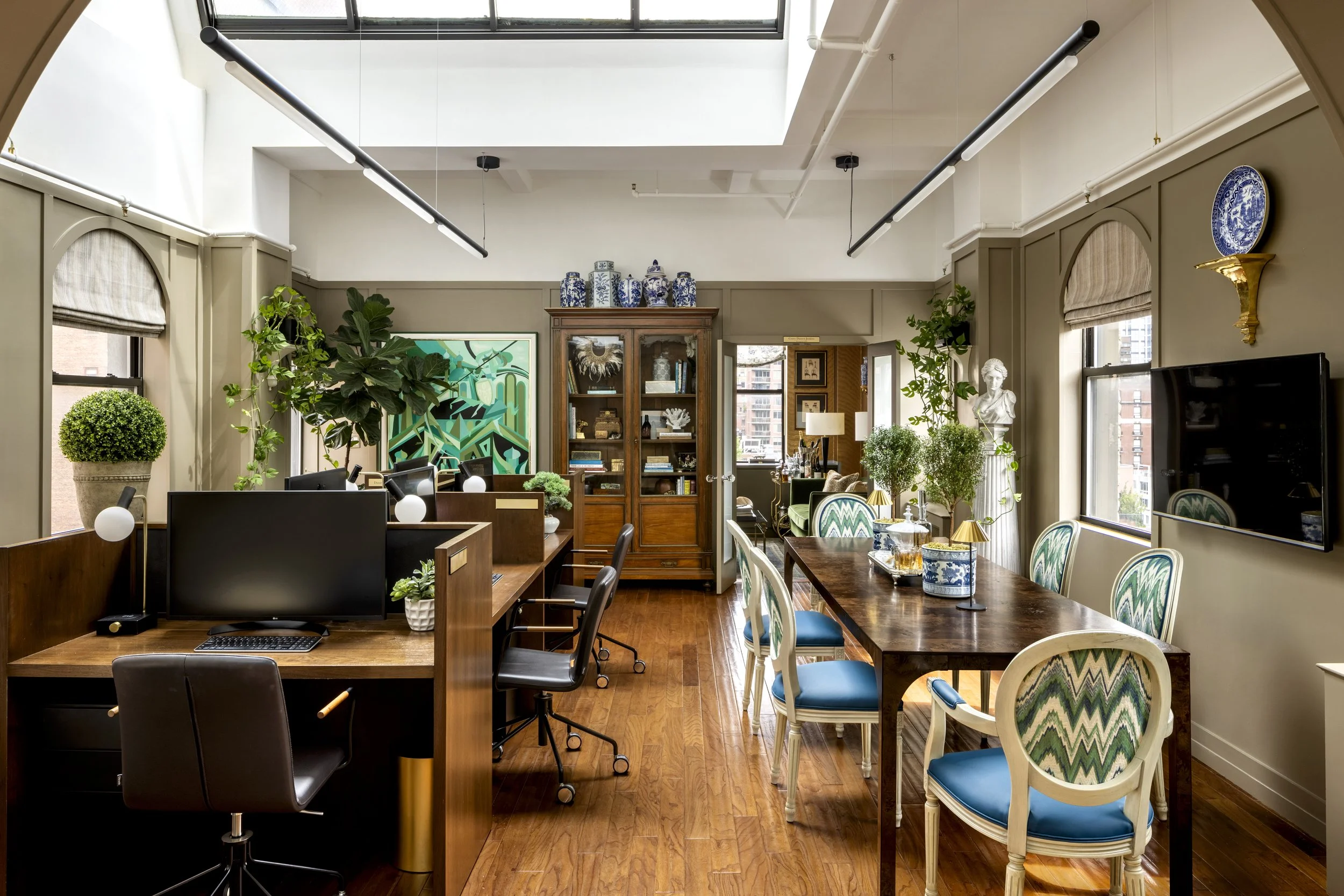 Bright, stylish office space with wooden furniture, green plants, large windows, decorative blue and white ceramics, a bust sculpture, and artwork on the wall.
