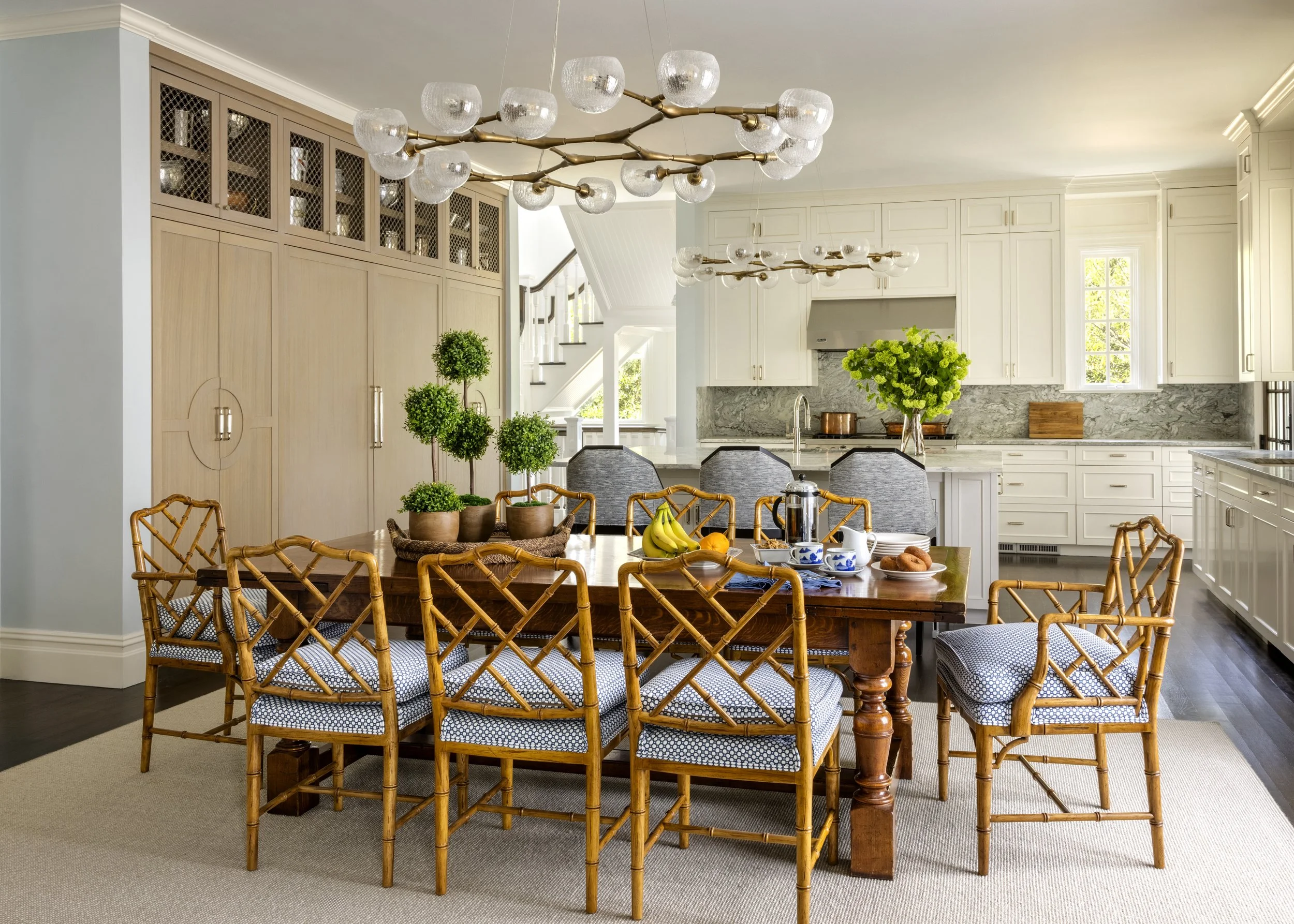 A dining room with a wooden table, surrounded by bamboo-style chairs with blue and white cushions, set with bananas, coffee cups, and pastries. The background features a white kitchen with cabinetry, a marble backsplash, and a window letting in natural light. Two modern chandeliers with glass globes hang from the ceiling.