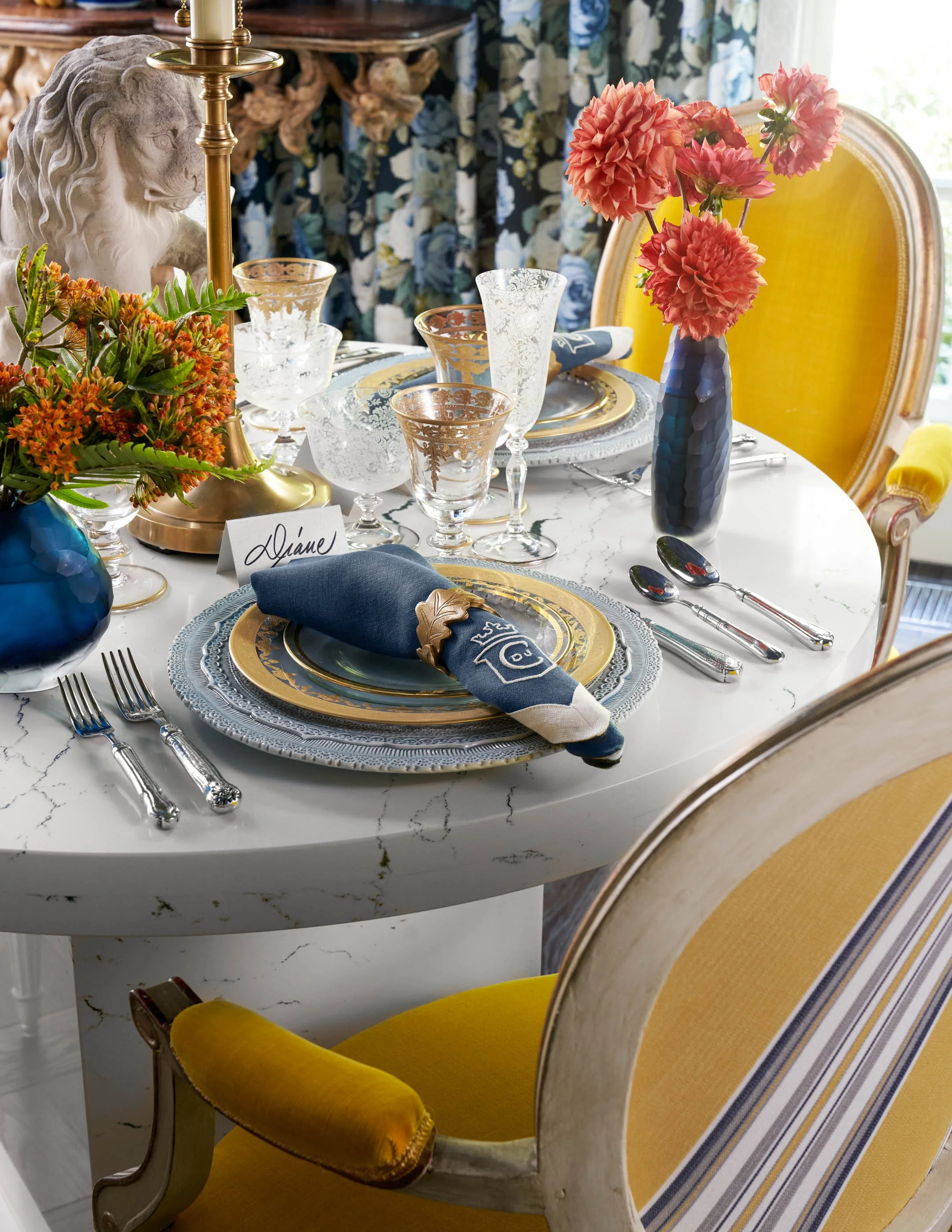 Elegant dining table with floral centerpiece, gold and crystal glassware, and china plates, set in a room with floral drapes and yellow upholstered chairs.