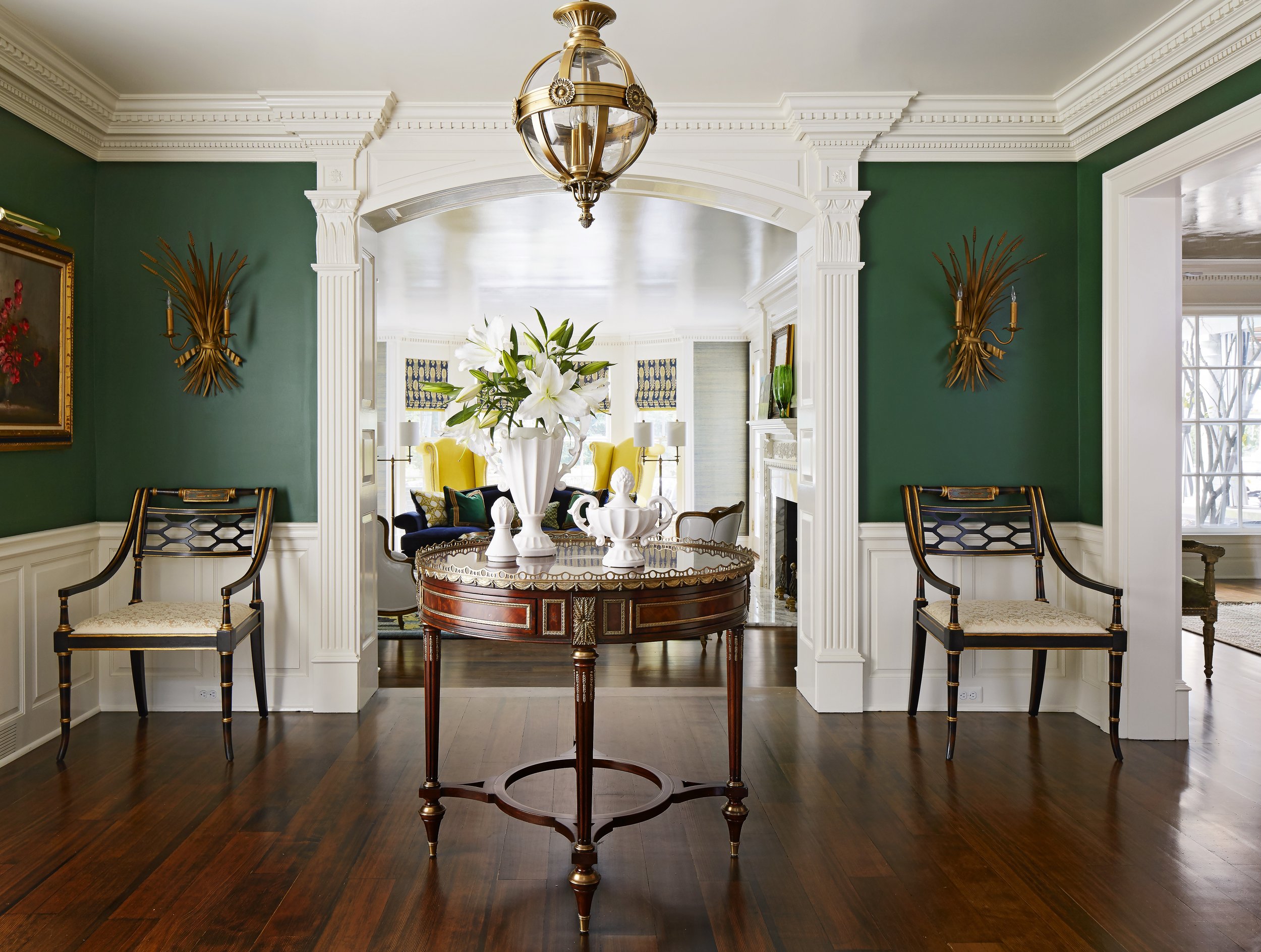 Elegant dining room with a round wooden table, a large floral centerpiece of white lilies, dark wood chairs, green walls, gold wall sconces, and a gold and glass chandelier.