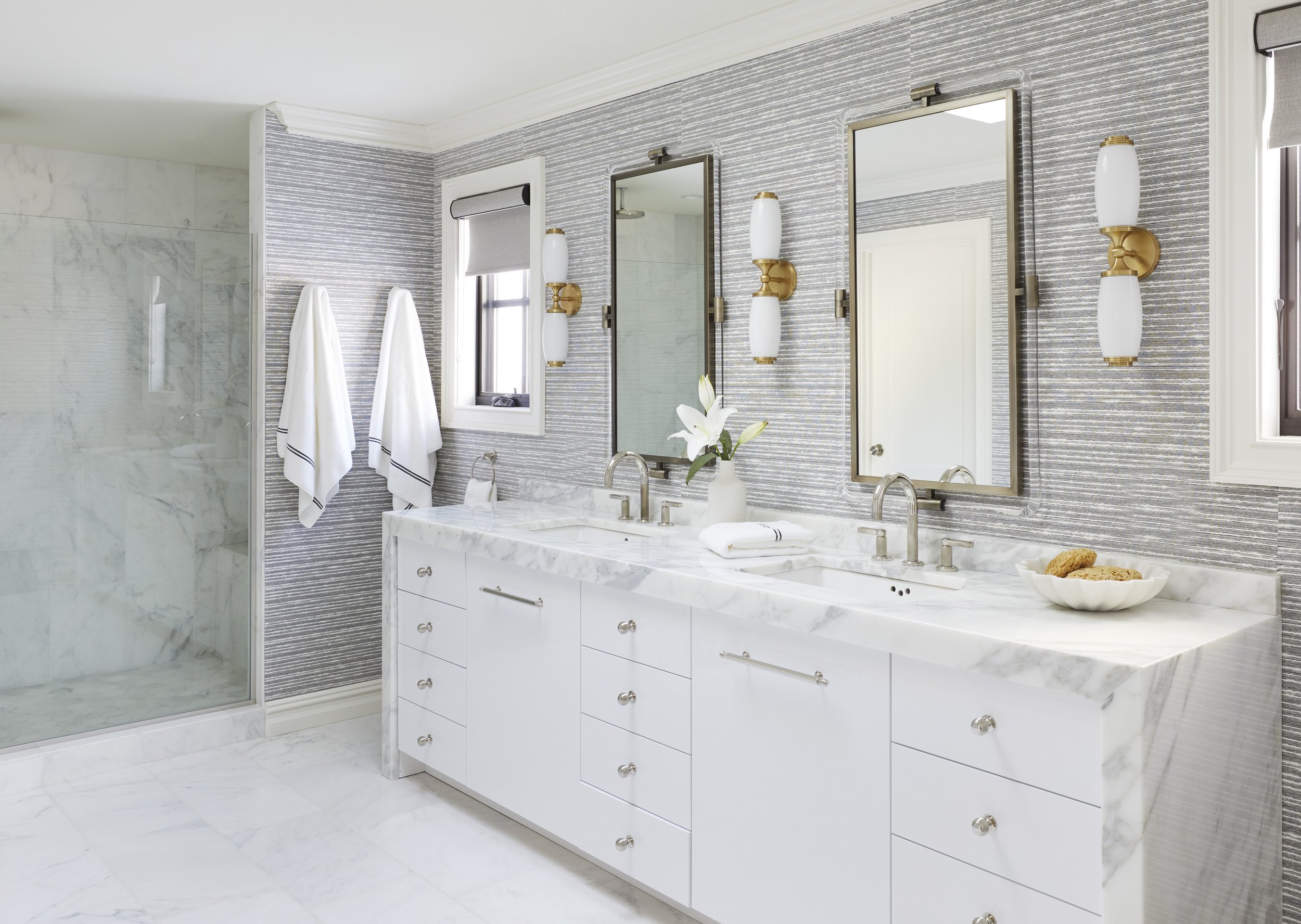 A modern bathroom featuring a marble-topped white vanity with drawers, large mirrors, gold wall sconces, and a marble shower enclosure with glass.