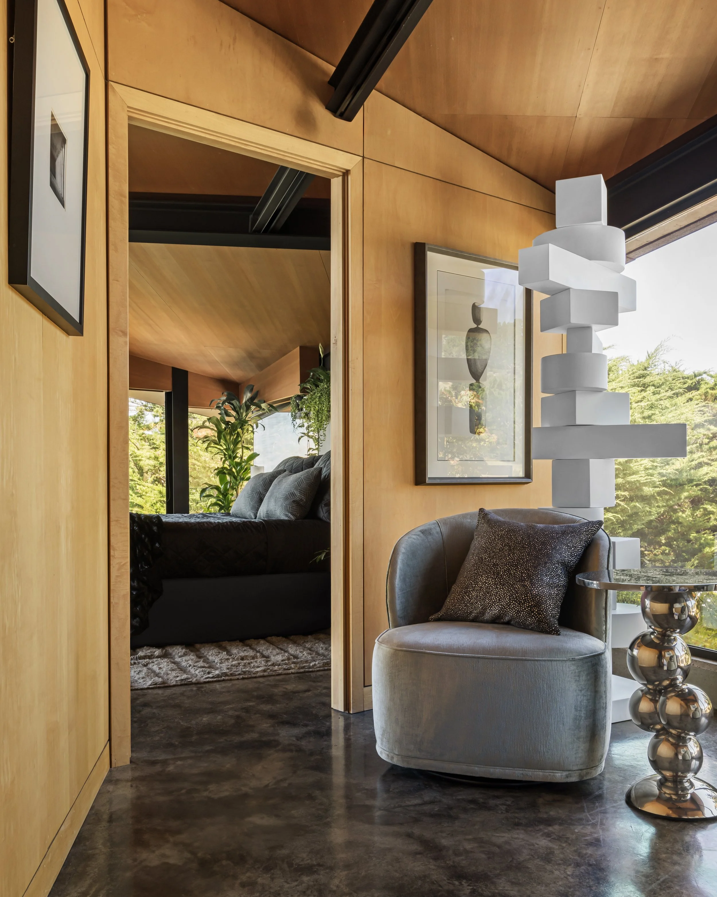 Modern living space with wood-paneled wall, framed artwork, white sculptural lamp, and a cozy gray armchair with a black and white patterned pillow, next to a small reflective metallic side table, with visible greenery outside through large windows.