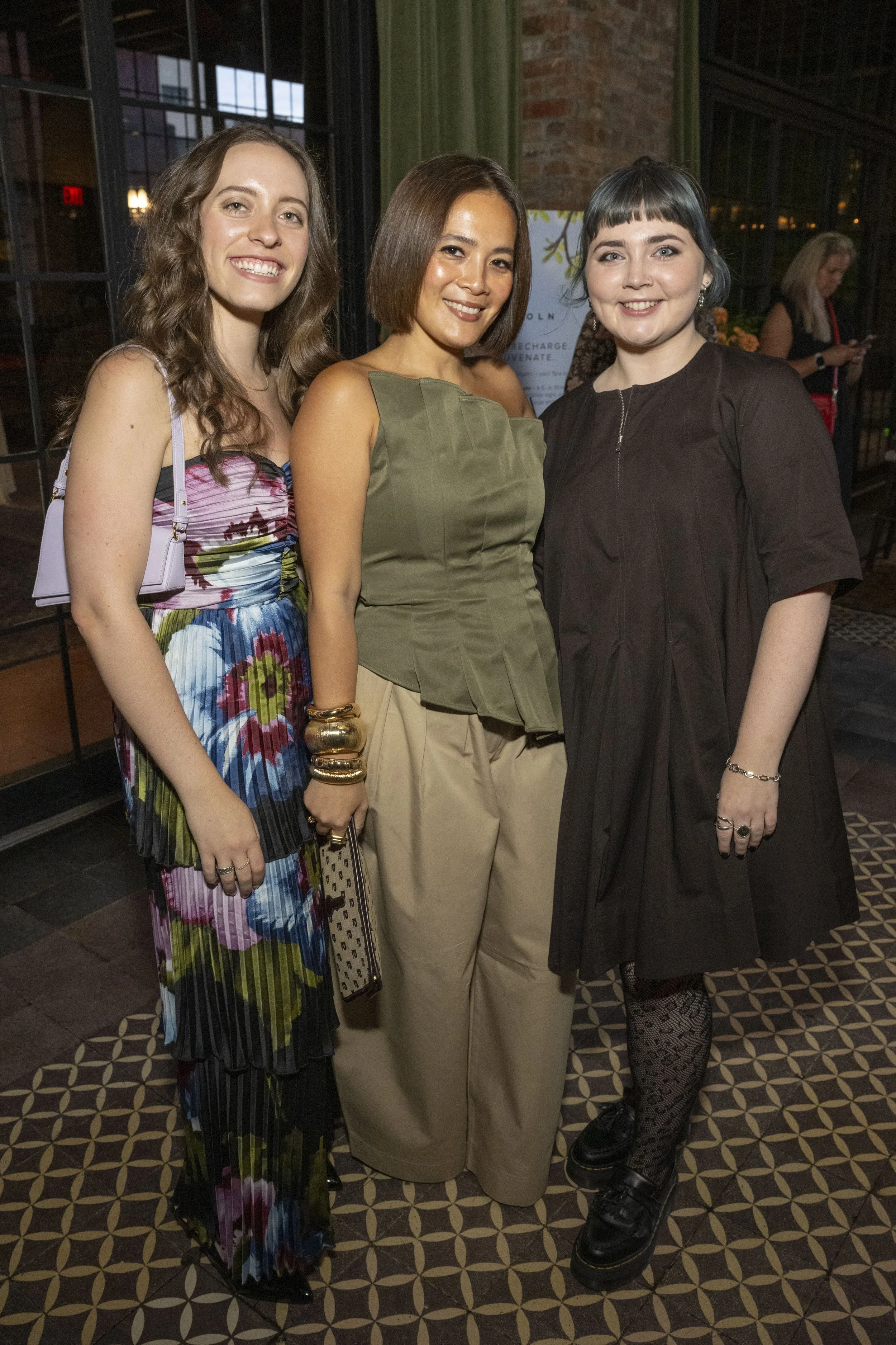 Three women standing side by side in a dimly lit indoor setting, smiling at the camera. The woman on the left has long, wavy brown hair and is wearing a colorful, sleeveless dress with a purple purse. The woman in the middle has a short, straight brown bob and is dressed in an olive-green top with beige pants. The woman on the right has short, dark hair with bangs and is wearing a black dress with fishnet stockings and black platform shoes.