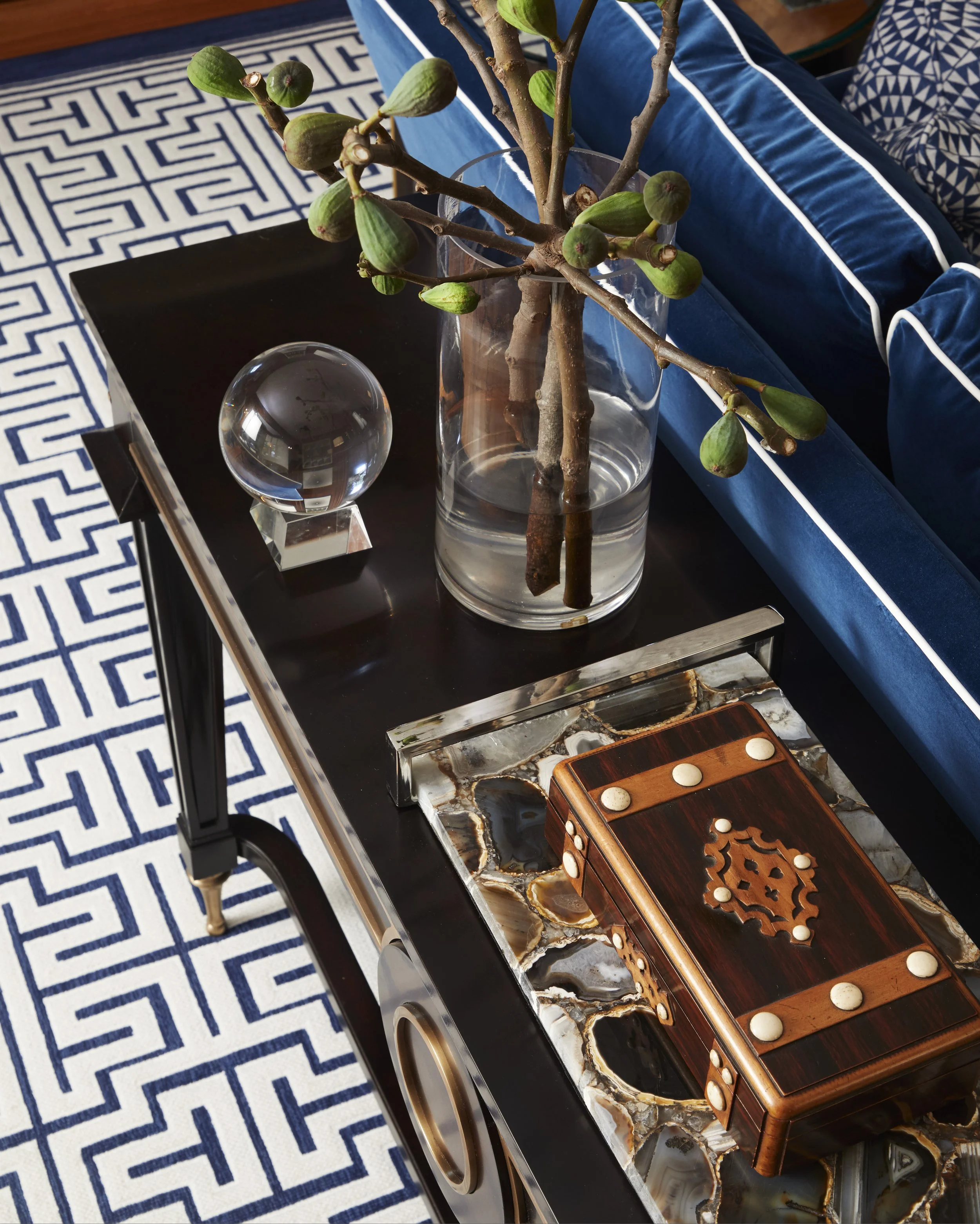 Decorative table with a glass vase holding a branch with green figs, a crystal ball on a black square base, and a wooden dominoes game board on a marble surface. The table is set against a background of a blue and white geometric patterned cloth and 