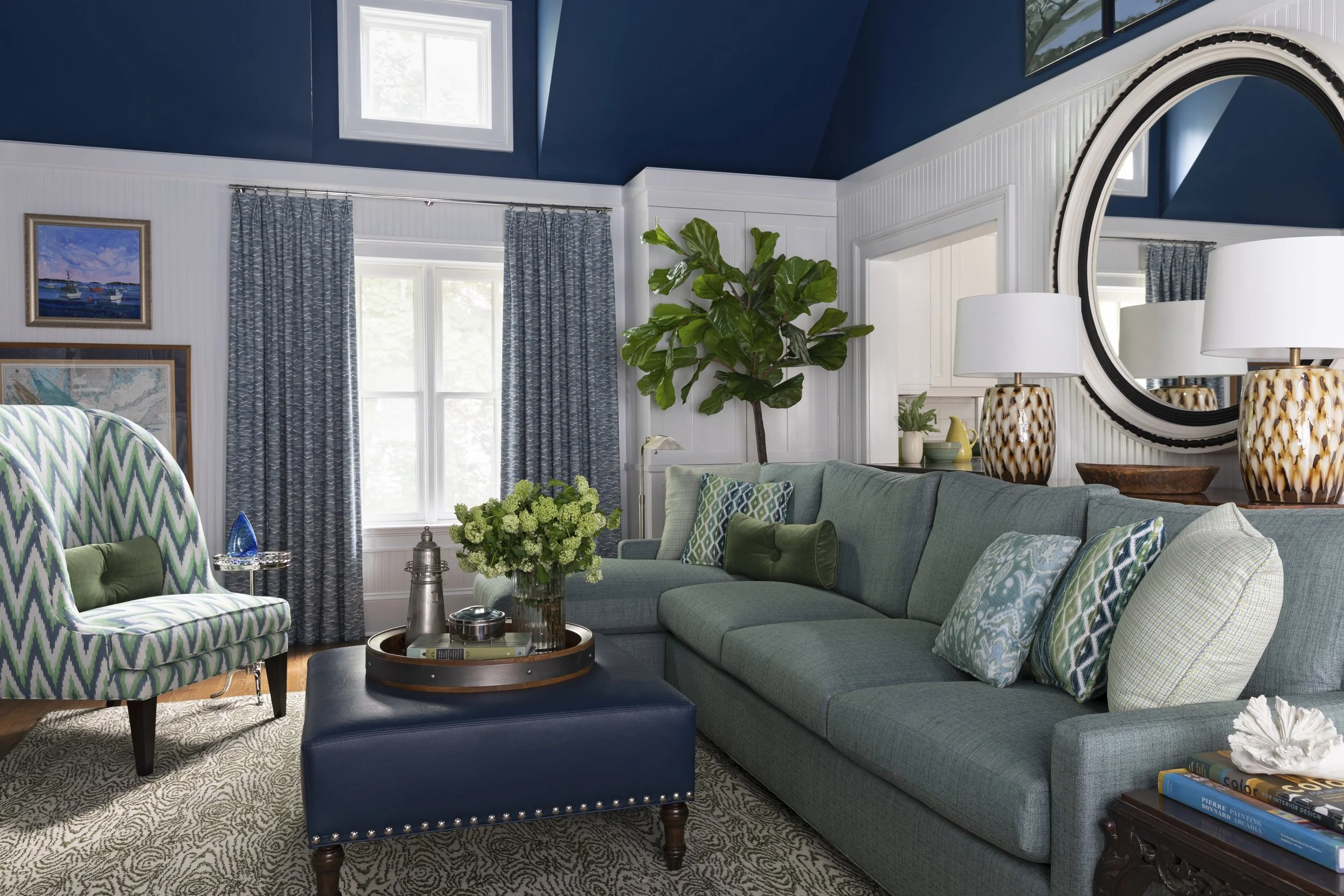 A cozy living room with a large light blue sectional sofa, patterned pillows, and a glass coffee table holding a floral arrangement and decorative items. Two matching lamps with patterned bases and white shades are on a side table. A large round mirr