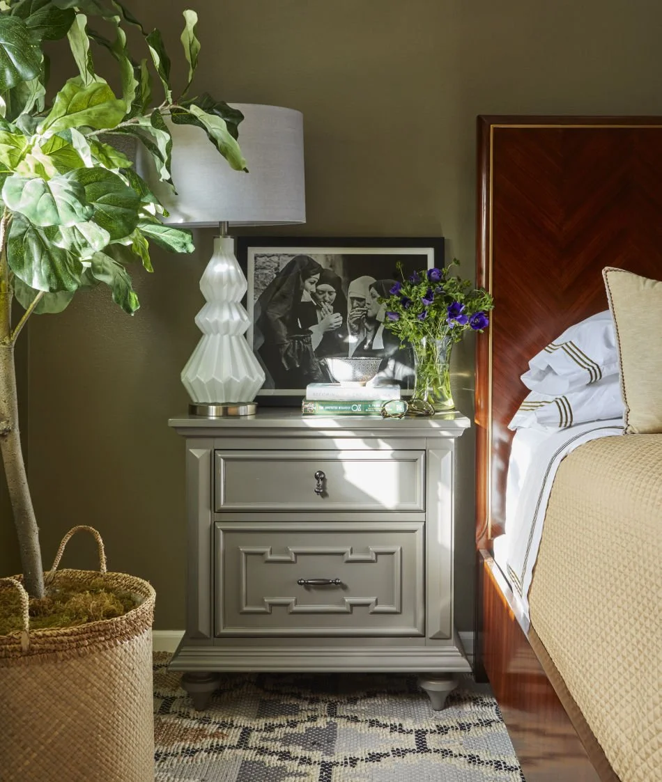 A bedroom with a nightstand holding a lamp, a framed black-and-white photo, a stack of books, and a vase with purple flowers. A bed with white bedding and a wooden headboard is adjacent, with part of a potted plant visible to the left.