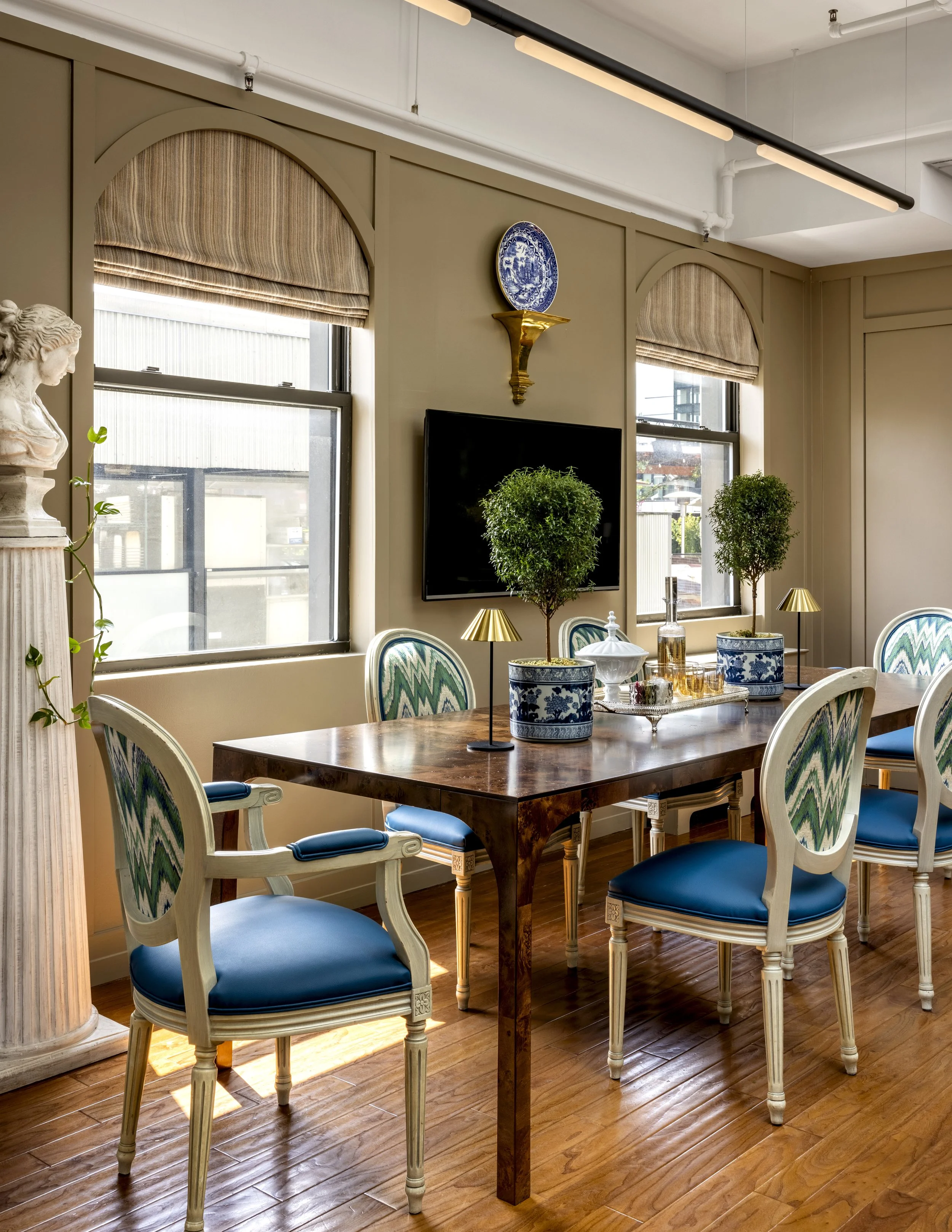 A dining room with a wooden table surrounded by white chairs that have blue upholstered seats and patterned backs. There are three potted plants on the table, each in blue and white decorative pots, and small gold lamps. The room has beige walls, two