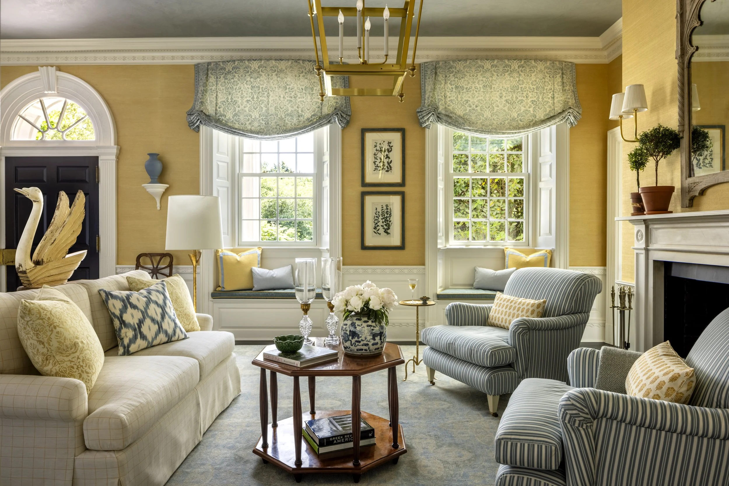 A living room with large windows, traditional decor, and a variety of couches and chairs. There is a wooden table with books and a flower vase in the center, and a gold chandelier hanging from the ceiling. The walls are yellow with blue curtains and 