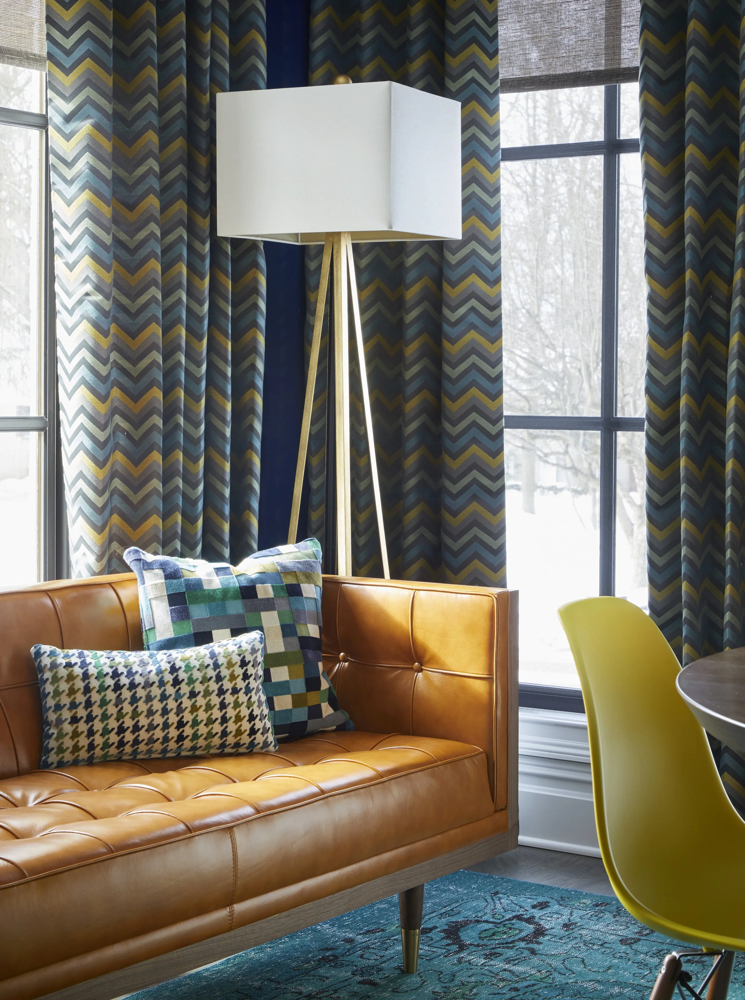 A cozy corner in a living room with a tan leather sofa and multicolored patterned cushions. Behind the sofa are large windows with blue, yellow, and green chevron-patterned curtains and sheer blinds. A tall, white and wood tripod floor lamp is next t