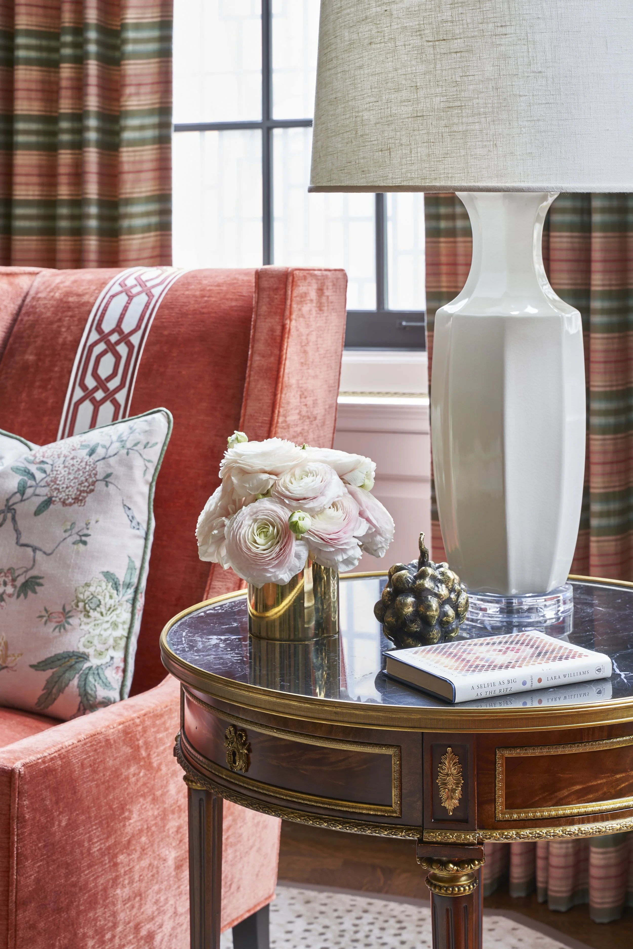 A side table with a black marble top and ornate gold trim, featuring a brass vase holding pale pink and white roses, two decorative gold-black pinecones, and a book titled 'A Selfie as Big as the Ritz' by Lara Williams. There is a coral-colored uphol