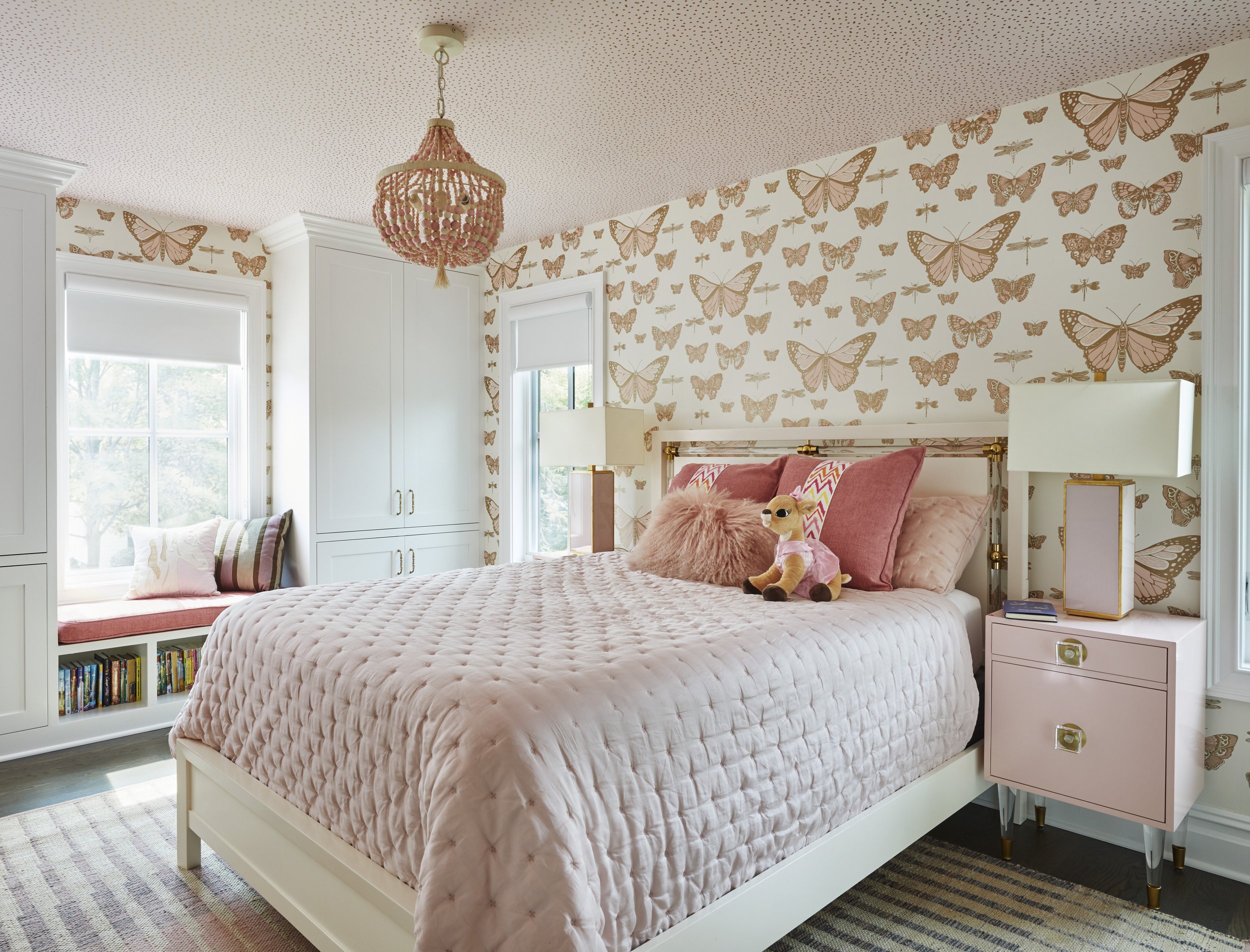 A bedroom with a pink bedspread, plush pillows, and stuffed animals, including a lion. The bedroom has white walls with wallpaper featuring butterfly and dragonfly patterns, white window frames, and built-in white cabinets. There is a pink bedside ta