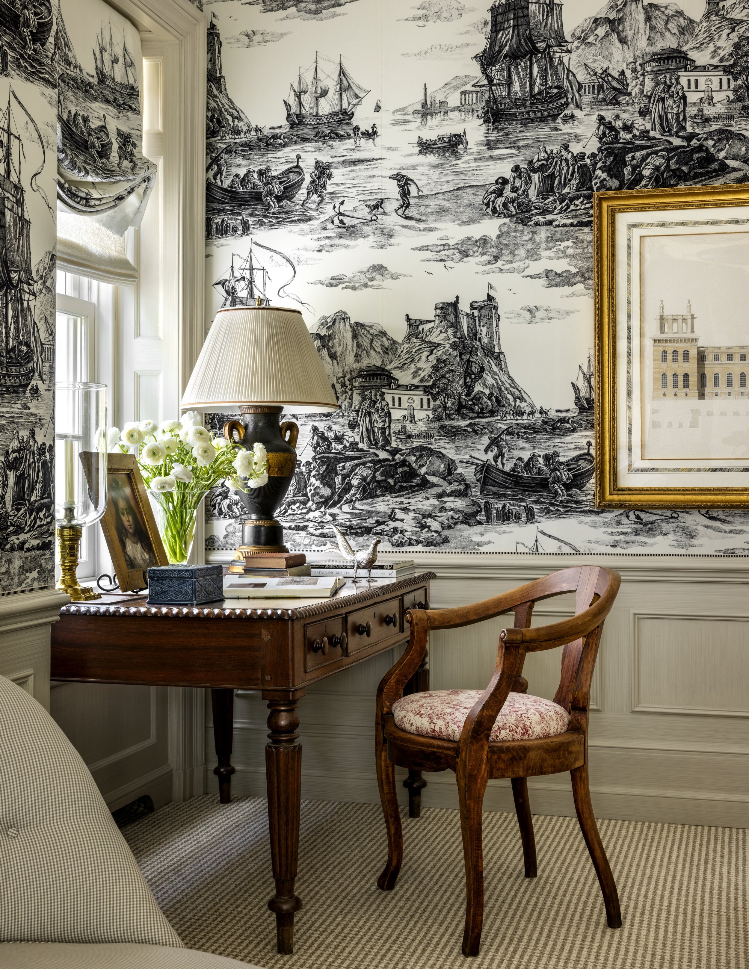 Interior of a room with a large maritime-themed mural, a wooden desk with a floral-lined lampshade, a vase of white flowers, a framed photograph, and a wooden chair with a pink-patterned cushion.