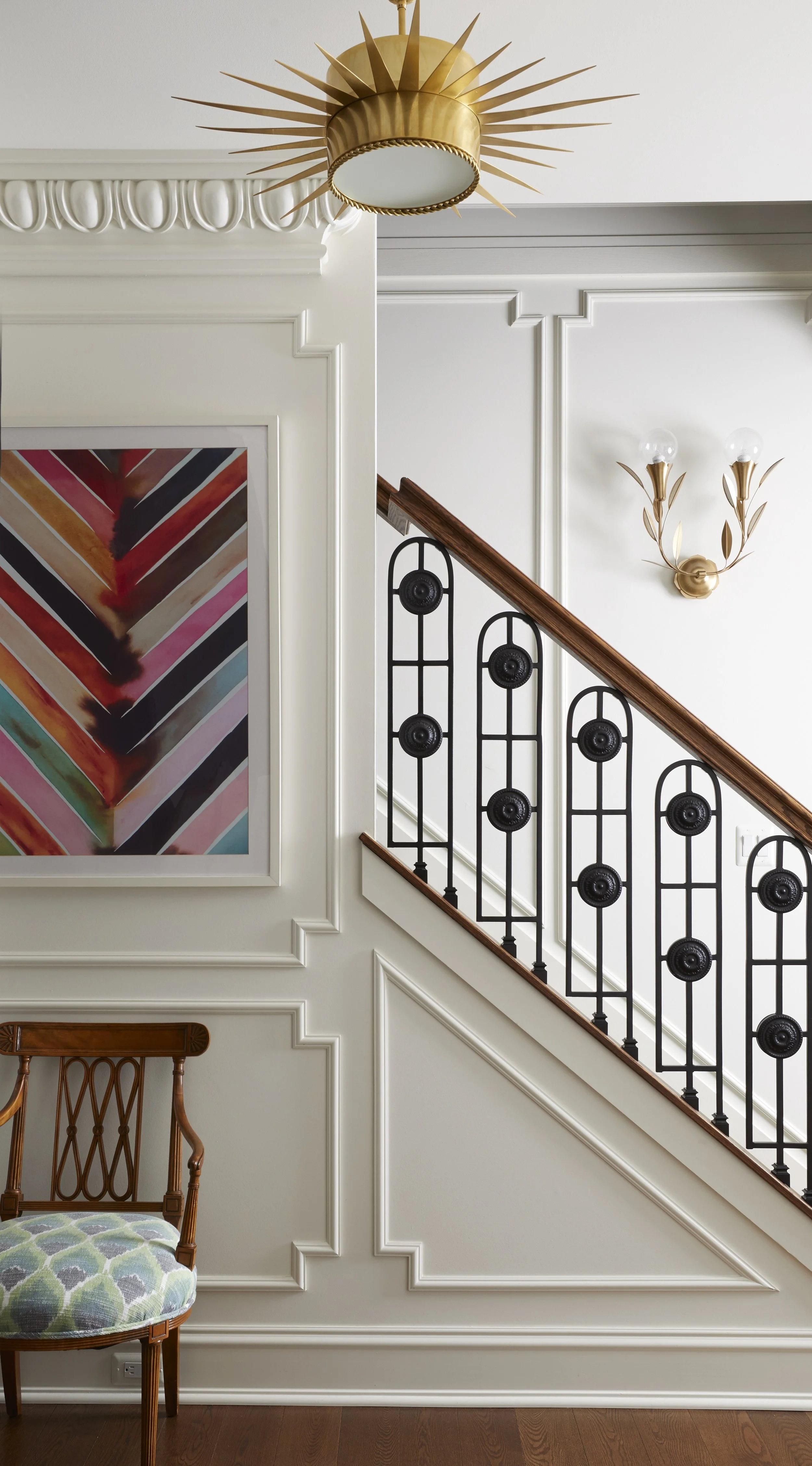 Interior of a house featuring a staircase with black decorative iron balusters, a wooden handrail, white paneled walls, a gold sunburst ceiling light, a wall-mounted light fixture with two exposed bulbs and gold accents, a colorful abstract painting,