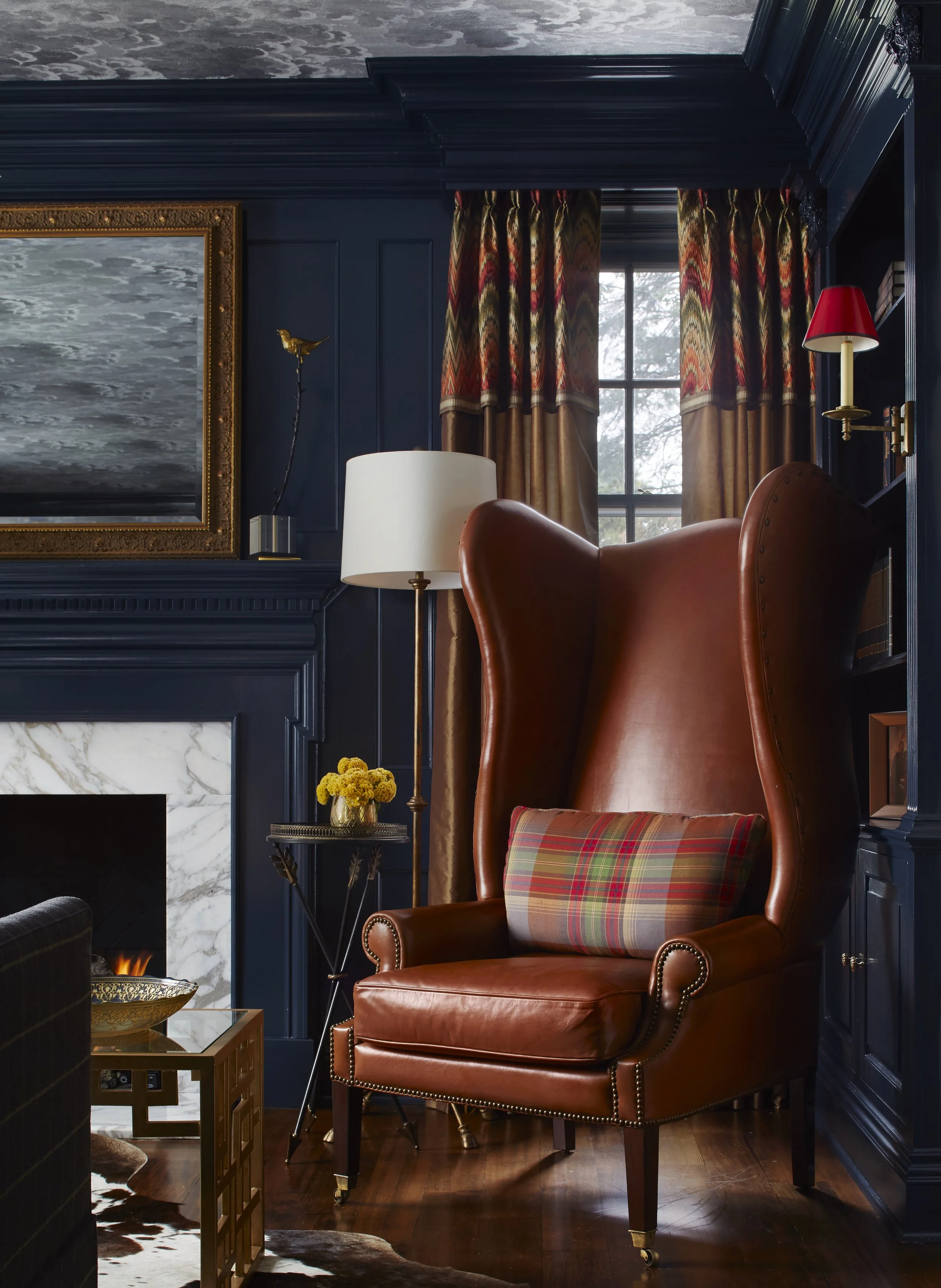 A cozy living room corner featuring a wingback leather chair with a plaid pillow, a side table with a yellow flower arrangement, a white lamp, and a decorative black and gold sculpture. There's a window with patterned curtains, and a fireplace with a