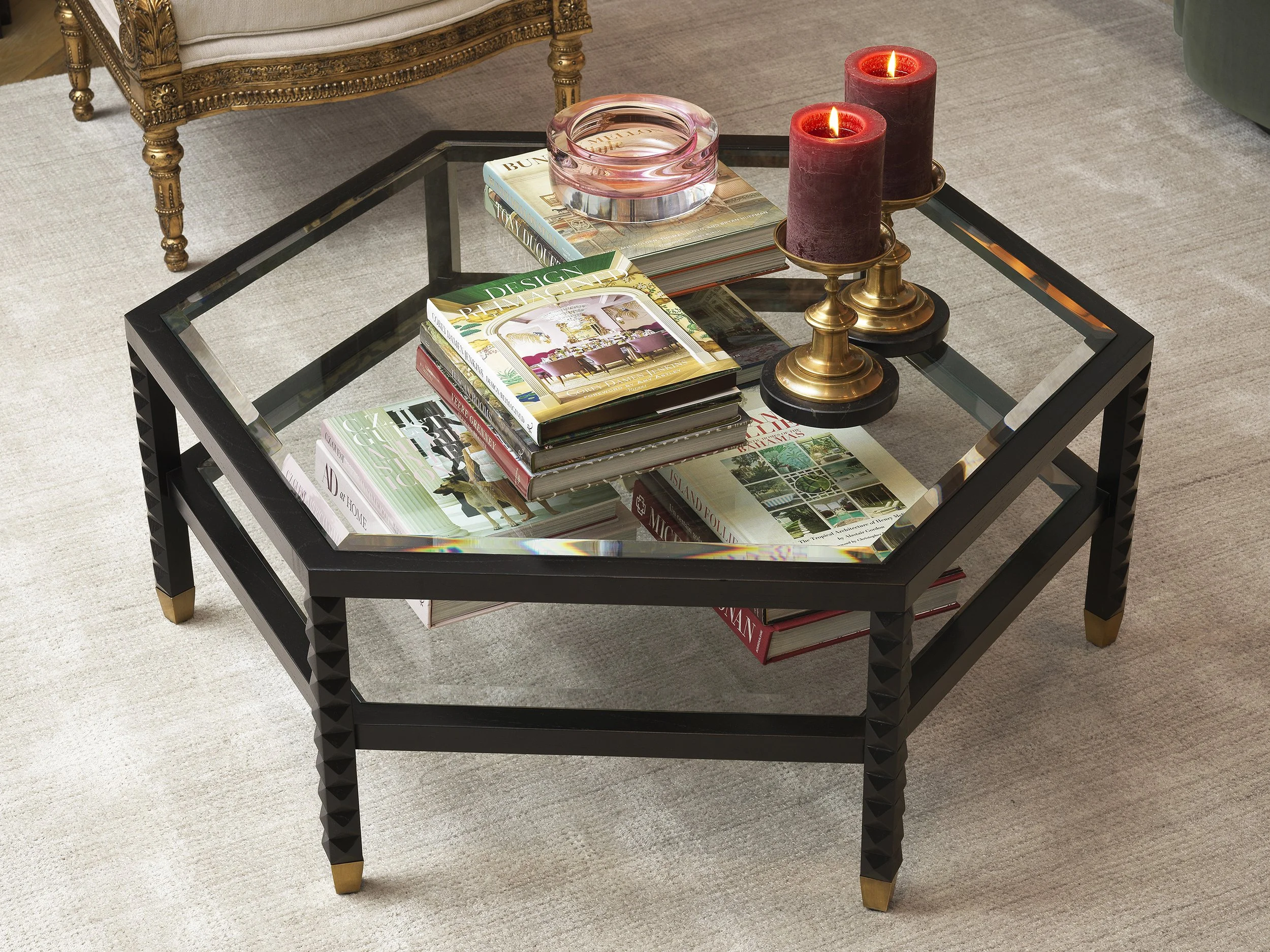 A glass-top coffee table with a glass sculpture, several stacked magazines, and two red candles on gold holders, situated in a room with a beige rug and ornate gold-finished furniture.