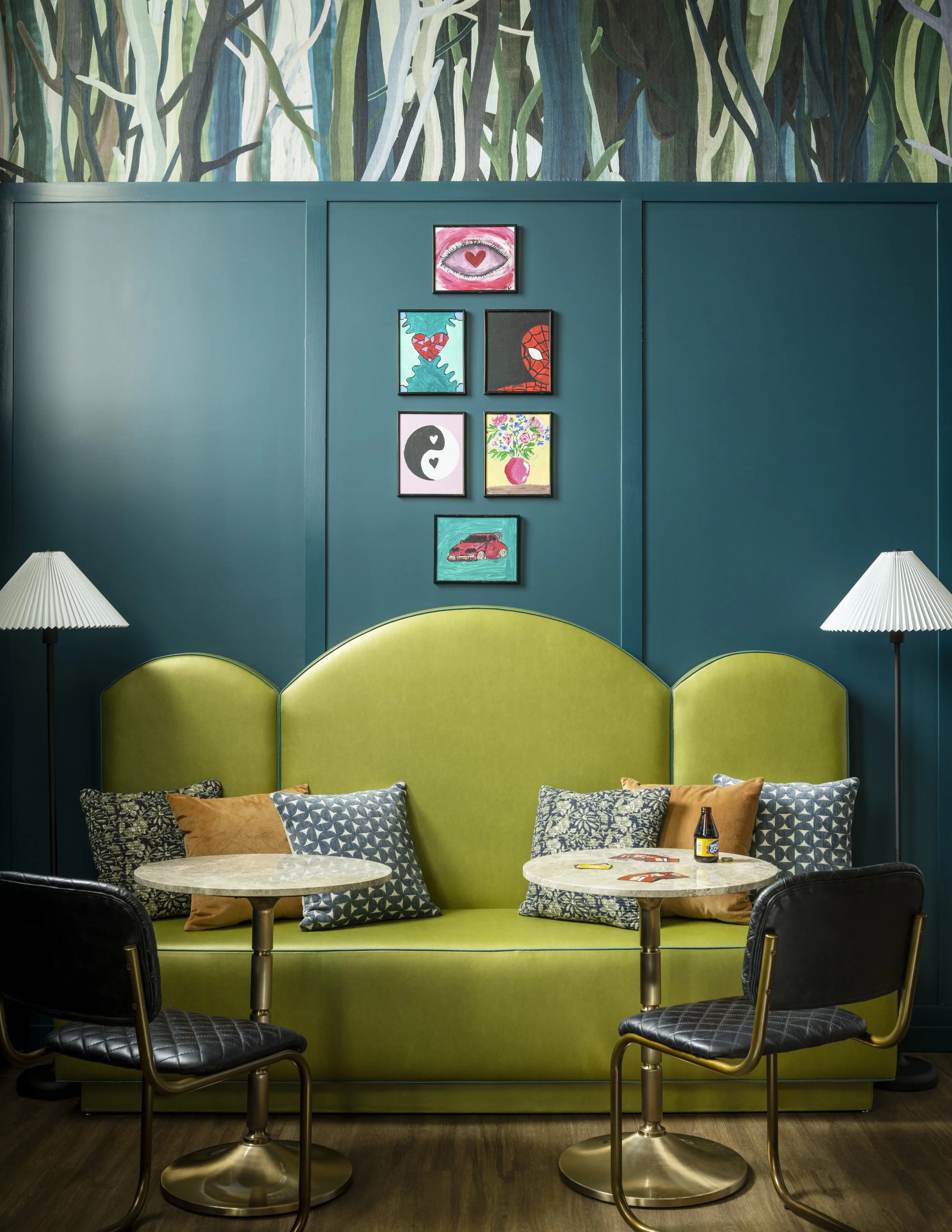A cozy dining area with two small round tables and black cushioned chairs, behind a vibrant green upholstered booth with decorative pillows. The wall behind features a teal color with a series of six framed artworks, including a pink eye with a heart