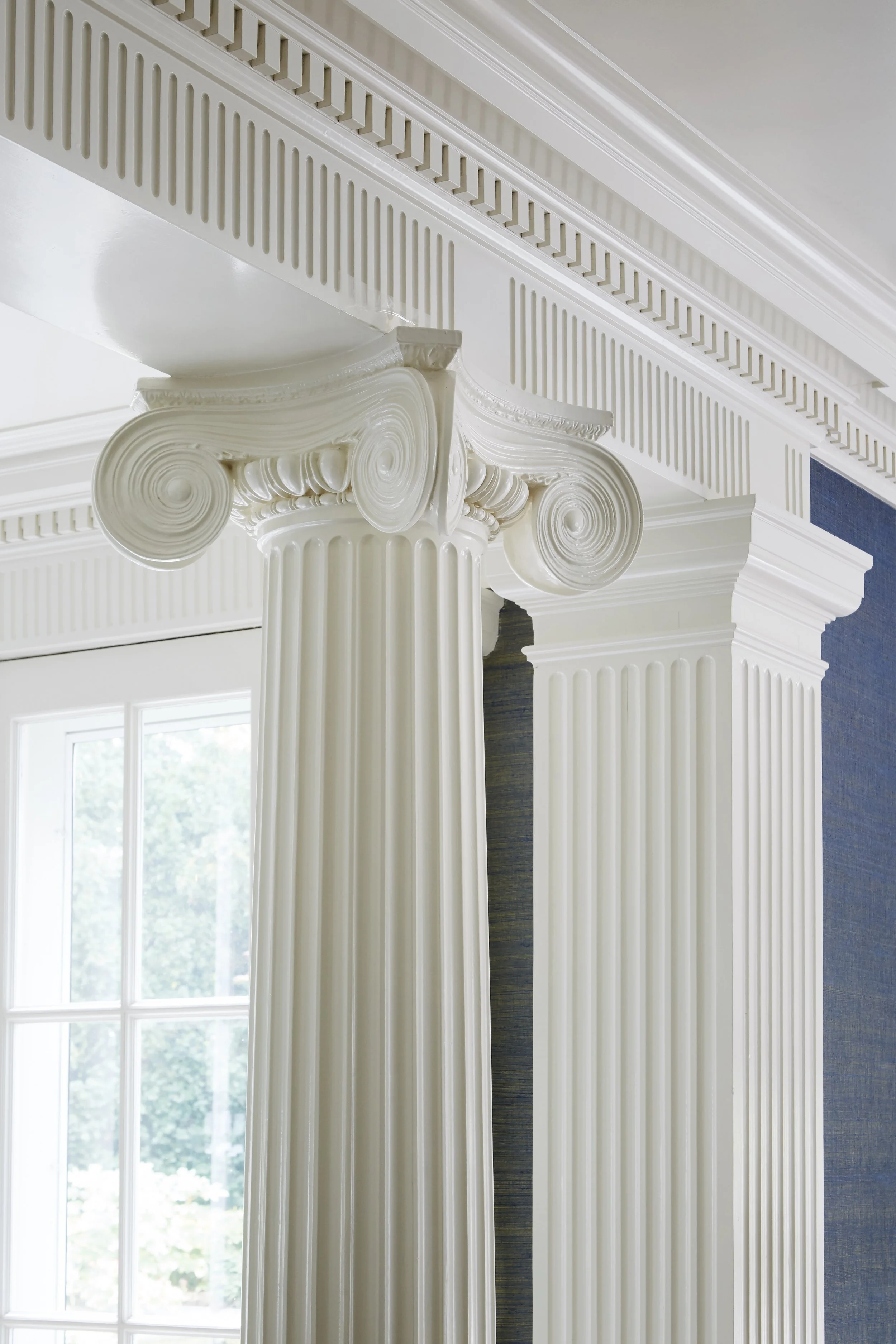 Close-up of white classical-style columns with ornate capitals, white crown molding, and a window in the background with trees outside.