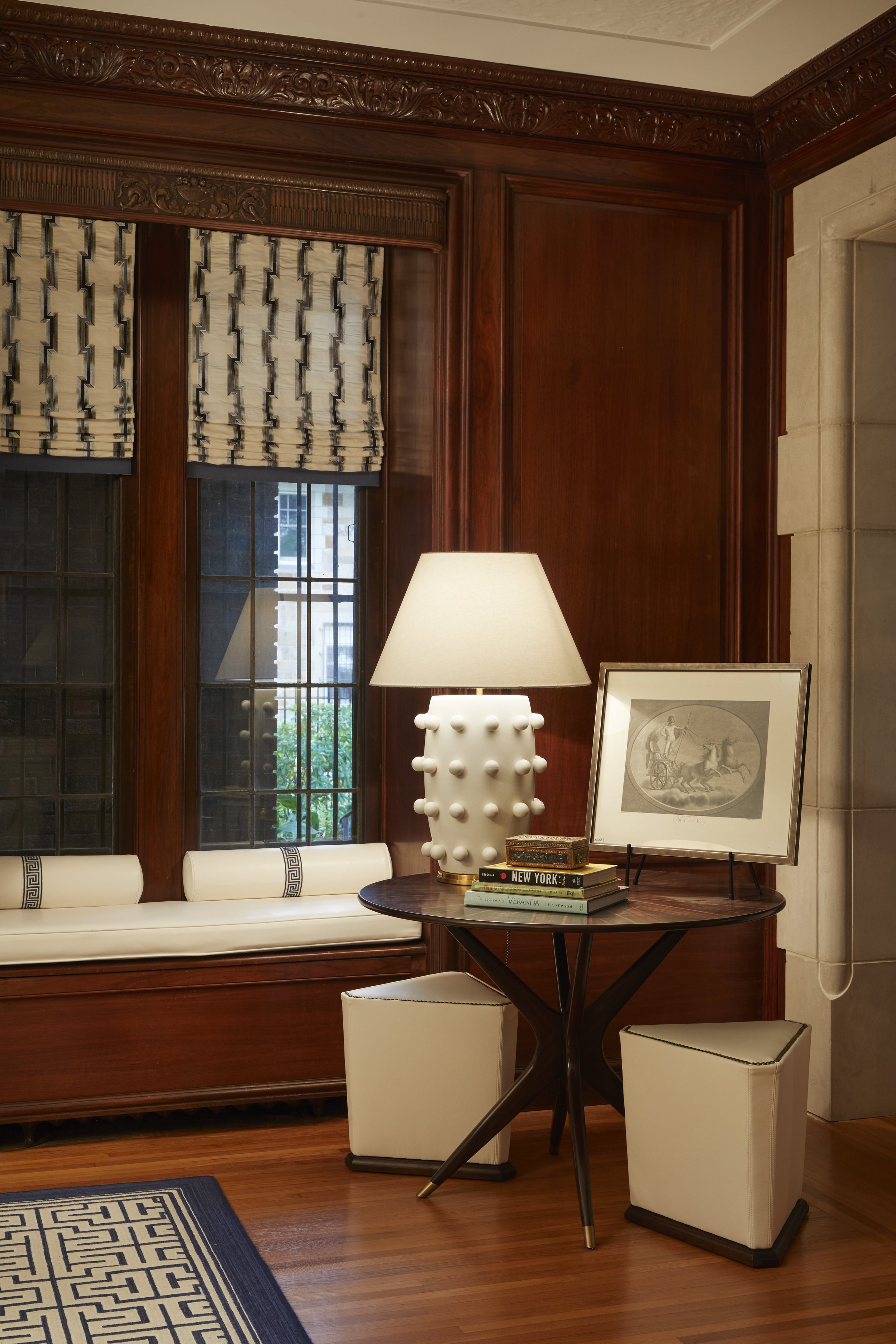 A room featuring dark wooden walls with ornate carvings, a window with patterned curtains, a white upholstered bench, a wooden round table with a stack of books and framed artwork, white stools, a white lamp with a textured base, and a blue and white