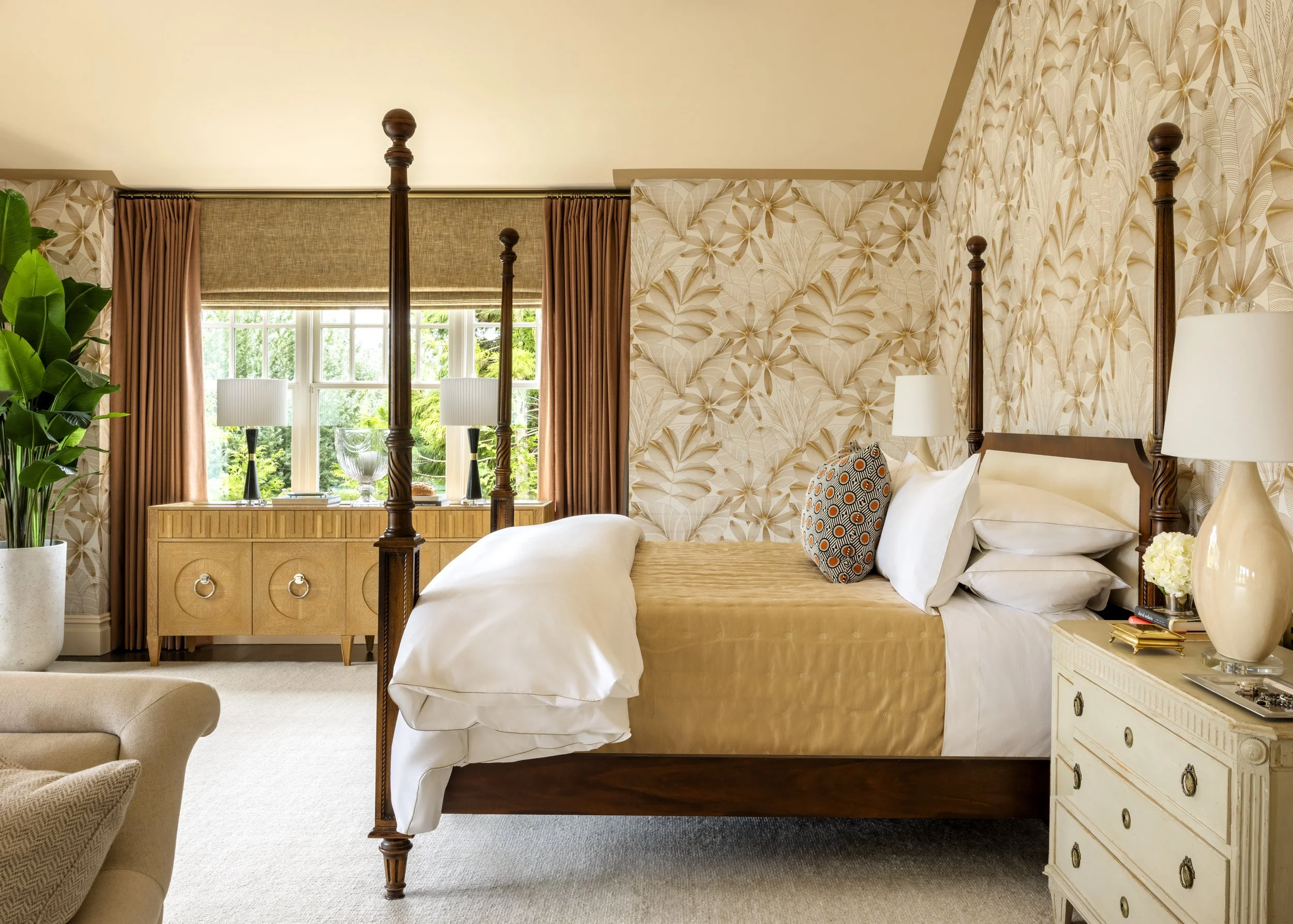 A cozy bedroom with beige and brown decor, a four-poster bed, patterned pillows, a white dresser, a lamp, greenery outside the window, and warm natural lighting.
