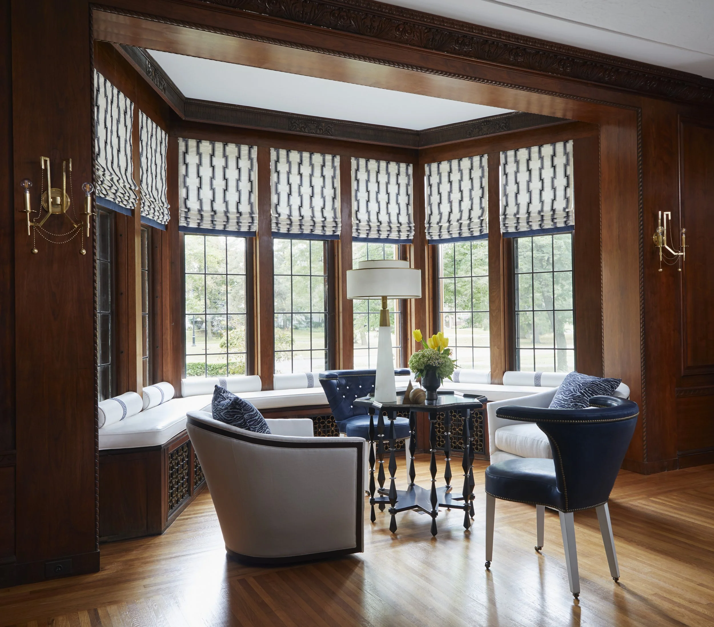A bright living room with large windows dressed with navy and white patterned Roman shades, dark wood crown molding, and a white sofa with navy and gray pillows, a round black table, a white table lamp, and a vase of yellow tulips.