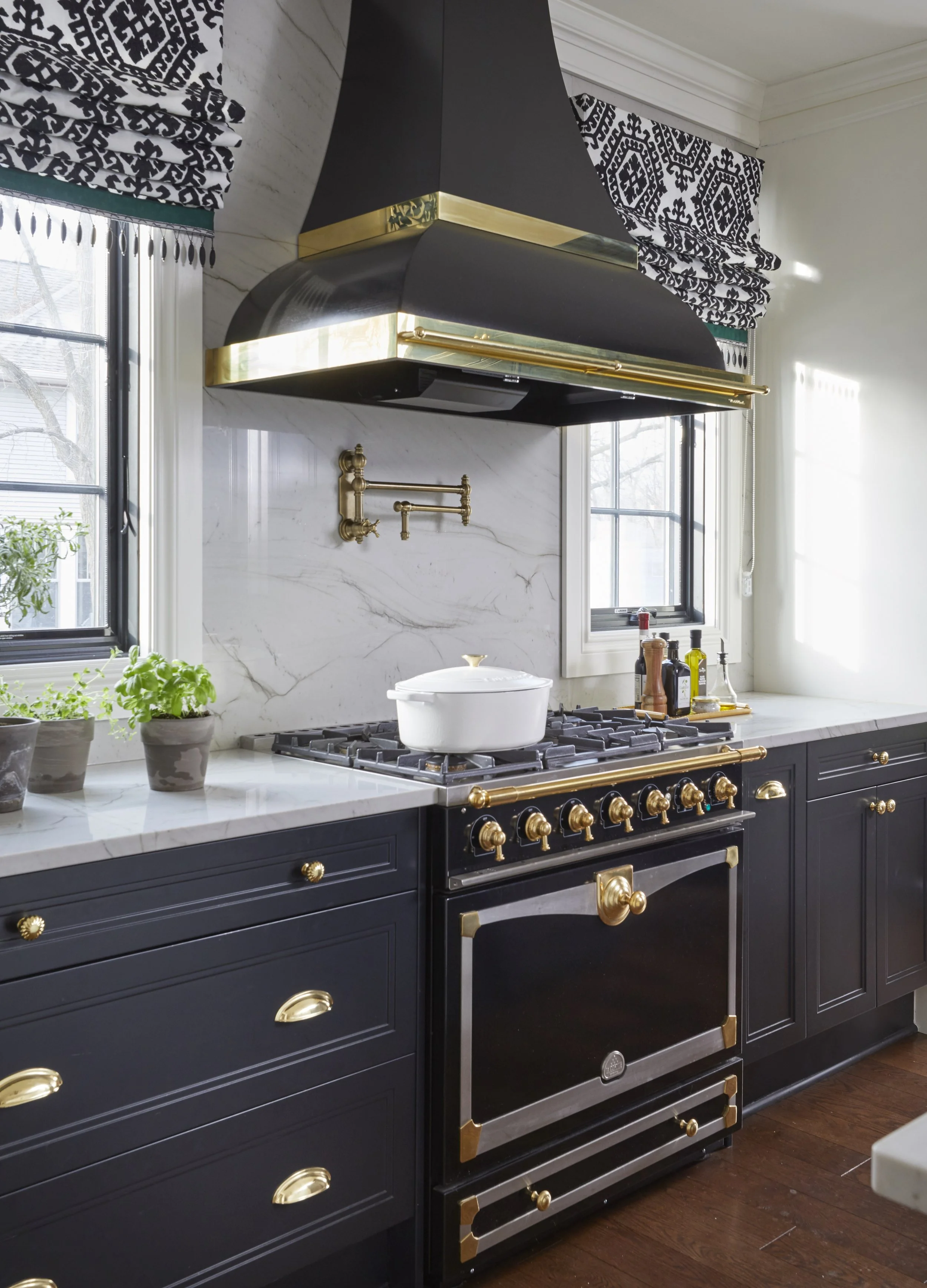 A kitchen with a black and gold stove and oven, marble countertops, a white ceramic pot on the stovetop, potted plants near the window, and black and white patterned curtains.