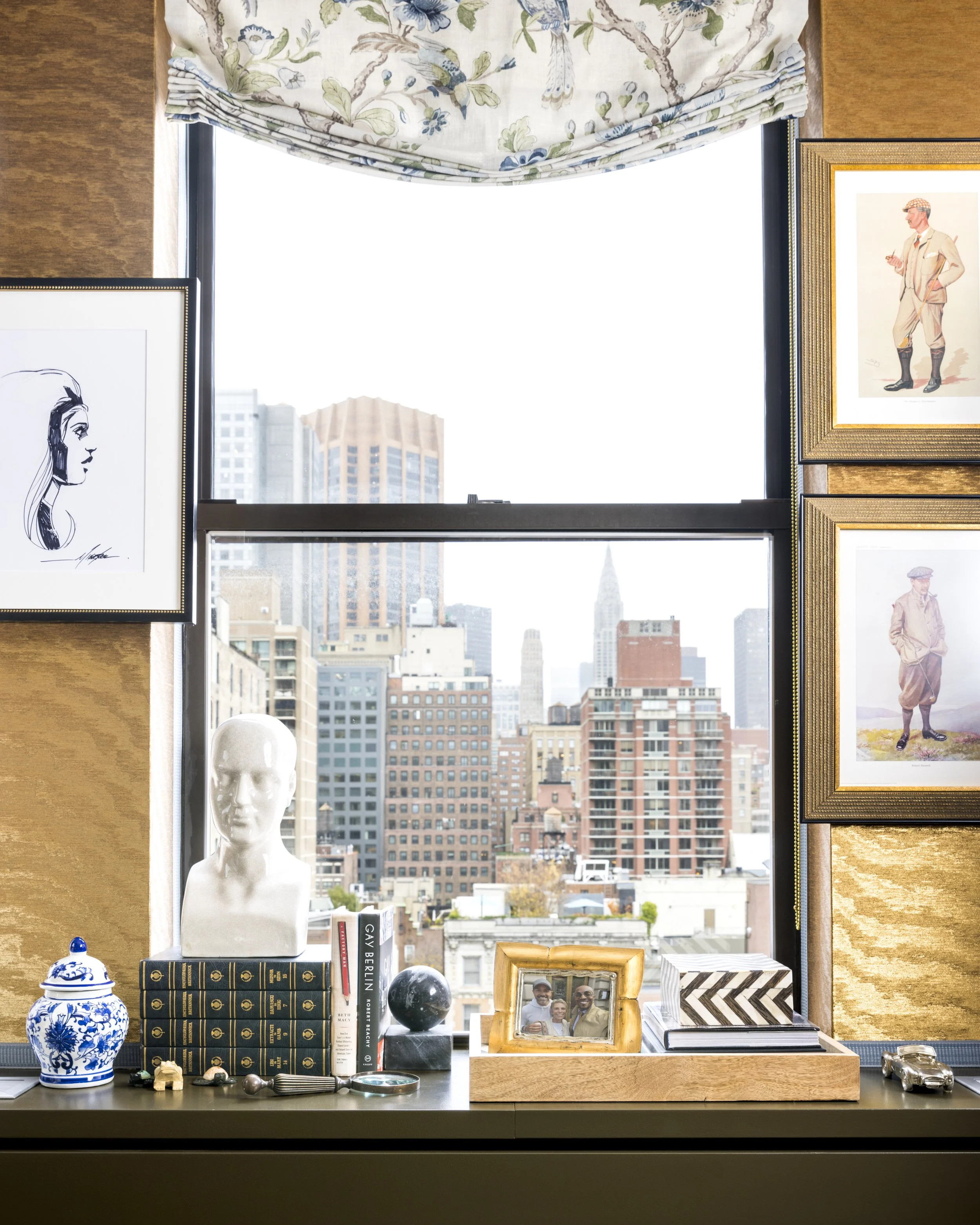 A windowsill display with a variety of objects including a white mannequin head, a blue and white vase, a stack of books, a framed photograph, decorative objects, and artwork, with a cityscape view in the background.