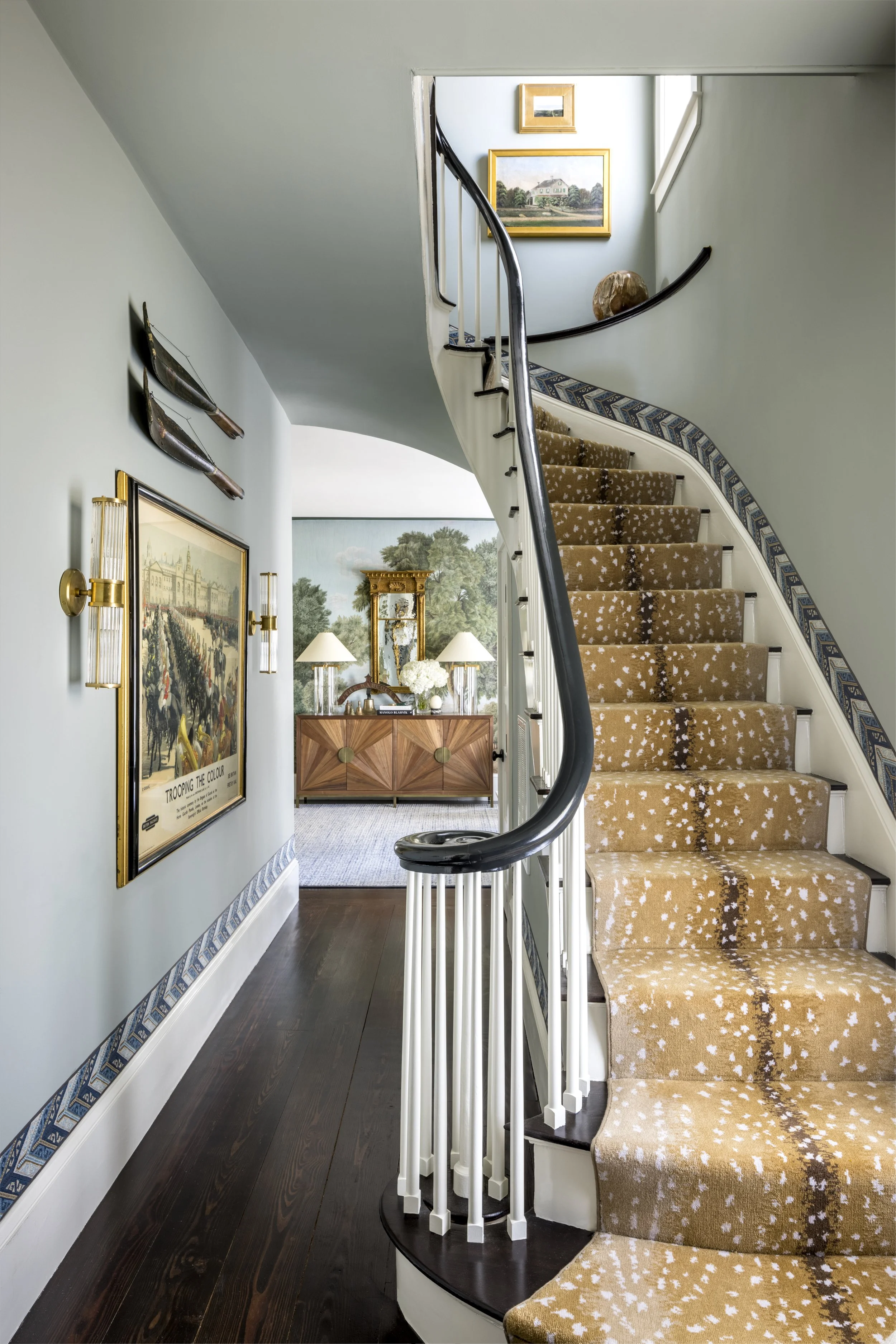 Interior view of a staircase with a dark wood floor, a curved staircase with beige carpet with white spots, and a black handrail. The walls are decorated with paintings, a mirror, and metal insect sculptures. There is a wooden console table with lamp