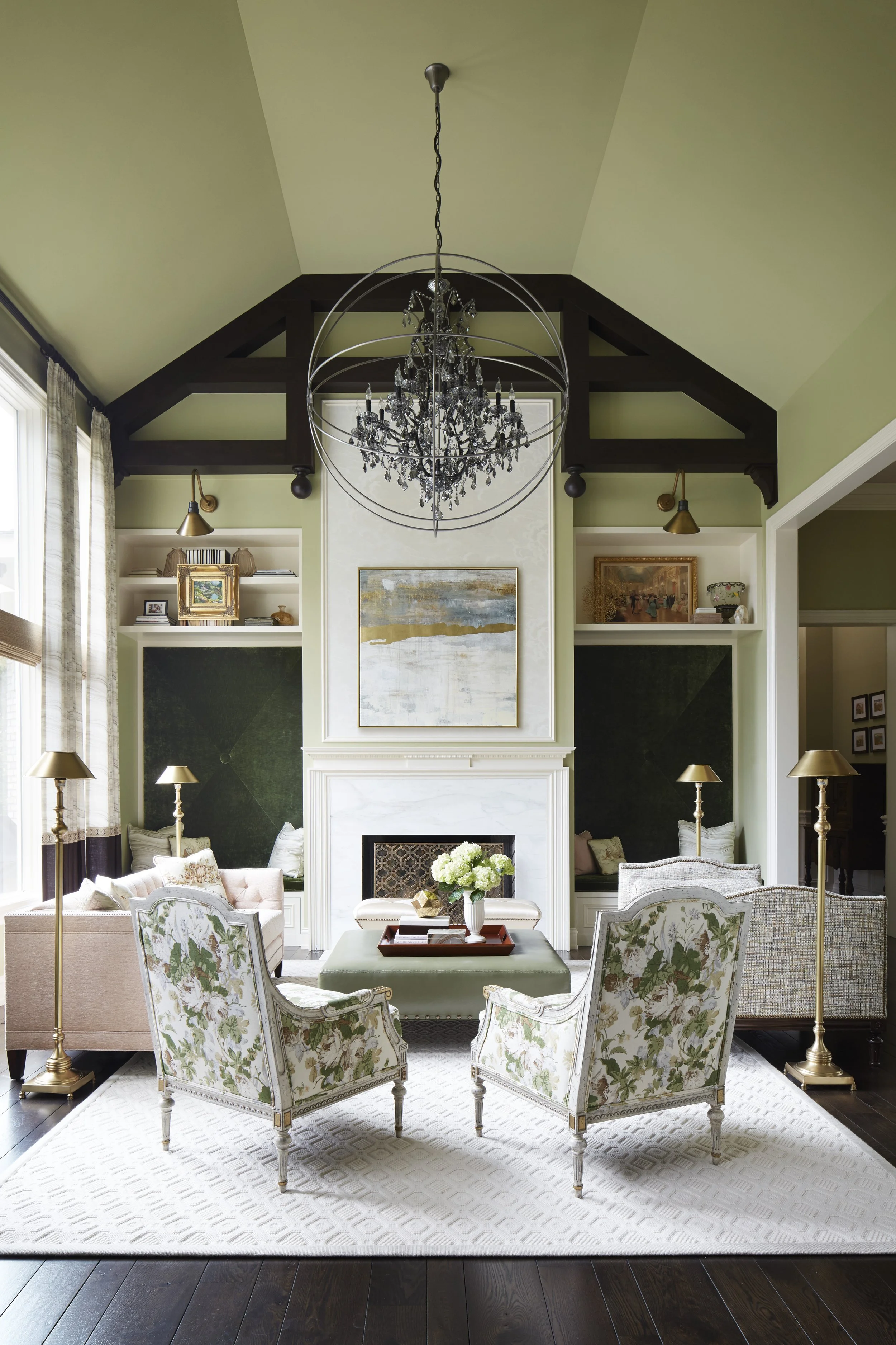 Elegant living room with a fireplace, floral armchairs, a chandelier, and a green accent wall with artwork and shelves