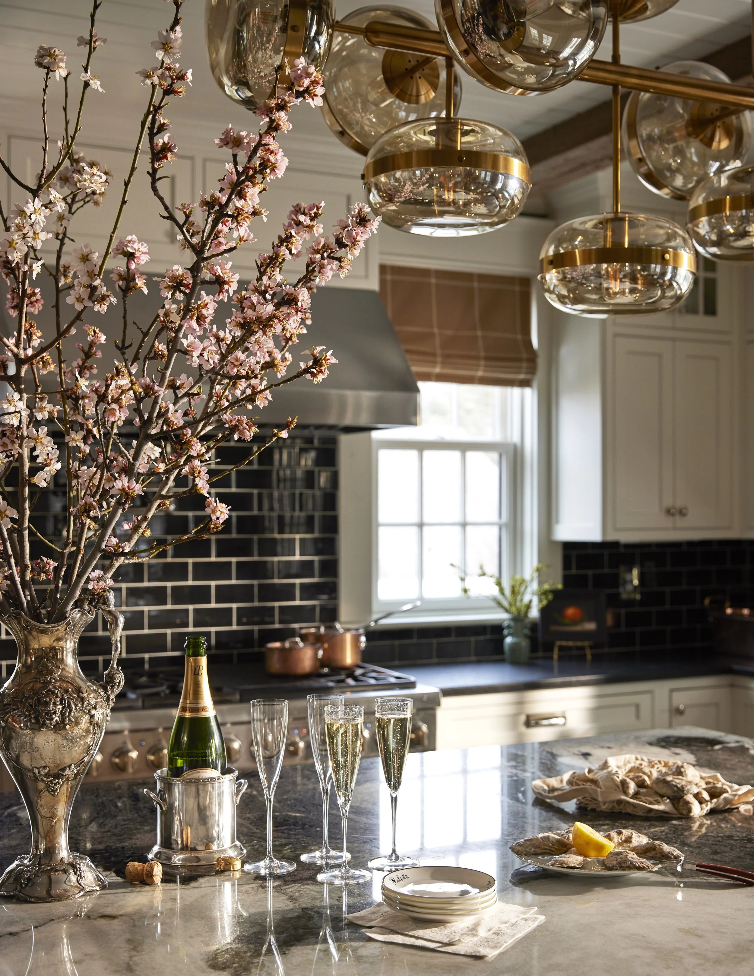 A kitchen counter decorated for celebration with a tall ornate silver vase holding pink cherry blossoms, champagne bottles, and several glasses filled with champagne. A tray of bread and lemon slice are on the counter, with a stack of small white pla