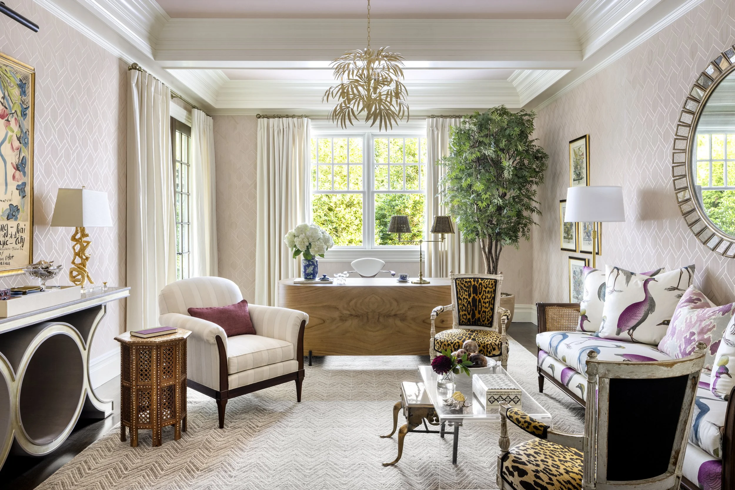Interior of a bright living room with large windows, white curtains, a chandelier, a large green potted plant, various chairs, a coffee table with decorative items, and artwork on the walls.