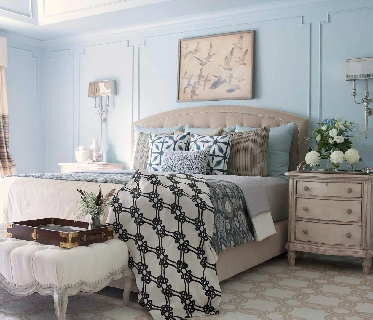 A cozy bedroom with a beige upholstered bed, multiple blue and patterned pillows, a abstract seagull painting above, white nightstands with vases and flowers, and a cream tufted bench at the foot of the bed. Light pastel blue walls and a decorative r