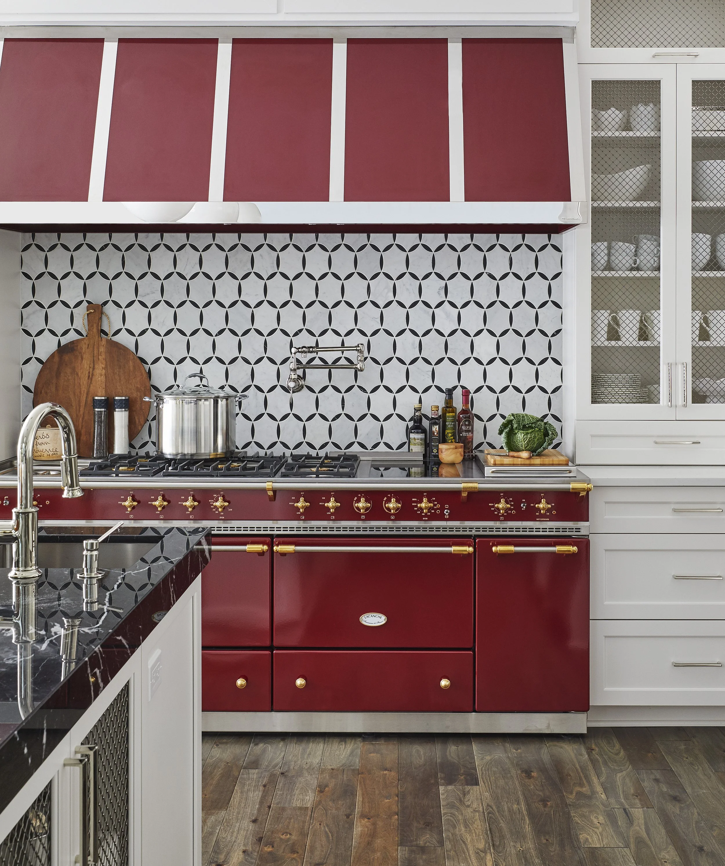 Stylish kitchen with black and white geometric backsplash, red cabinetry with gold hardware, black marble countertop, and open shelving with dishes.