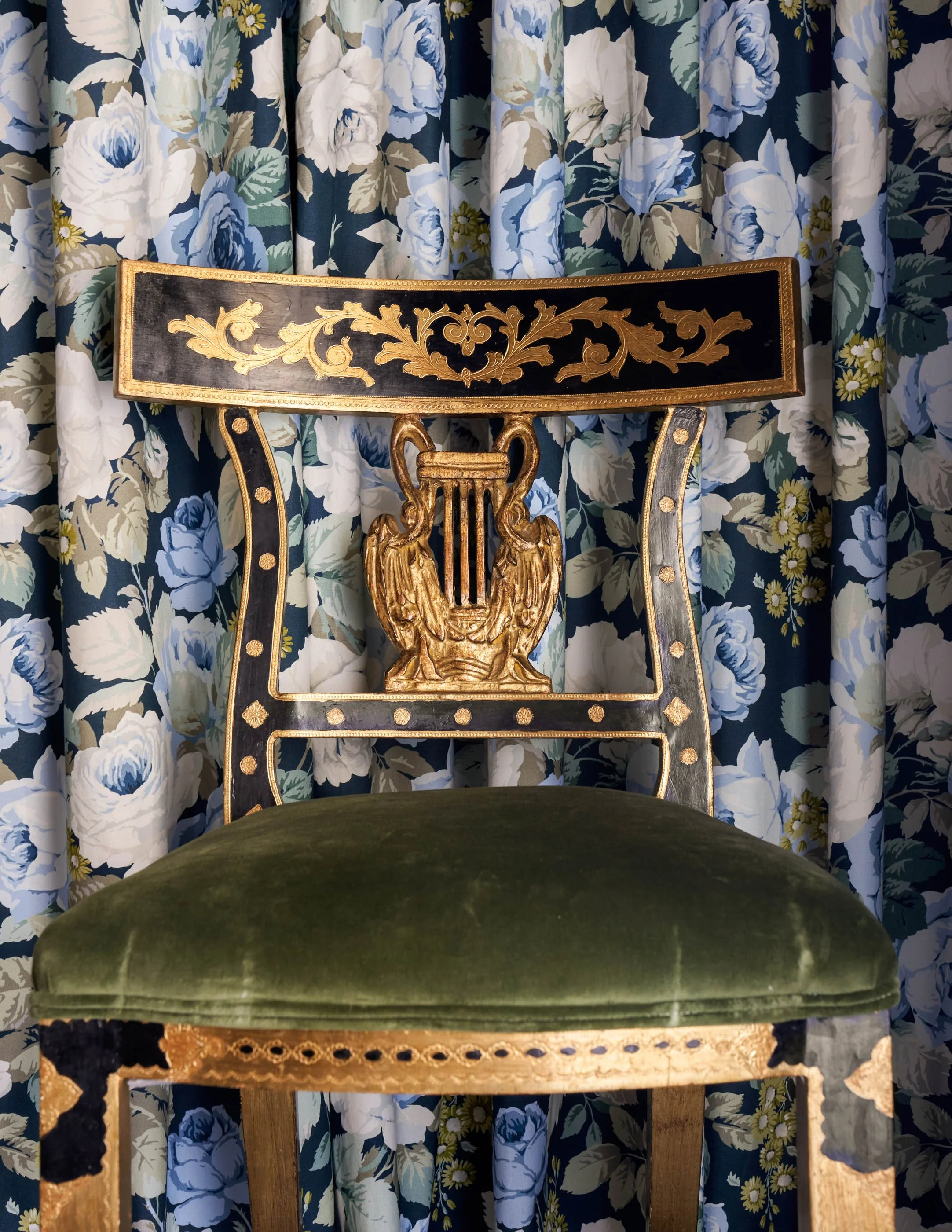 An ornate chair with a dark green velvet cushion, gold decorative trim, and a carved gold emblem on the backrest. The chair is positioned in front of floral curtains with a blue, white, and yellow flower pattern.