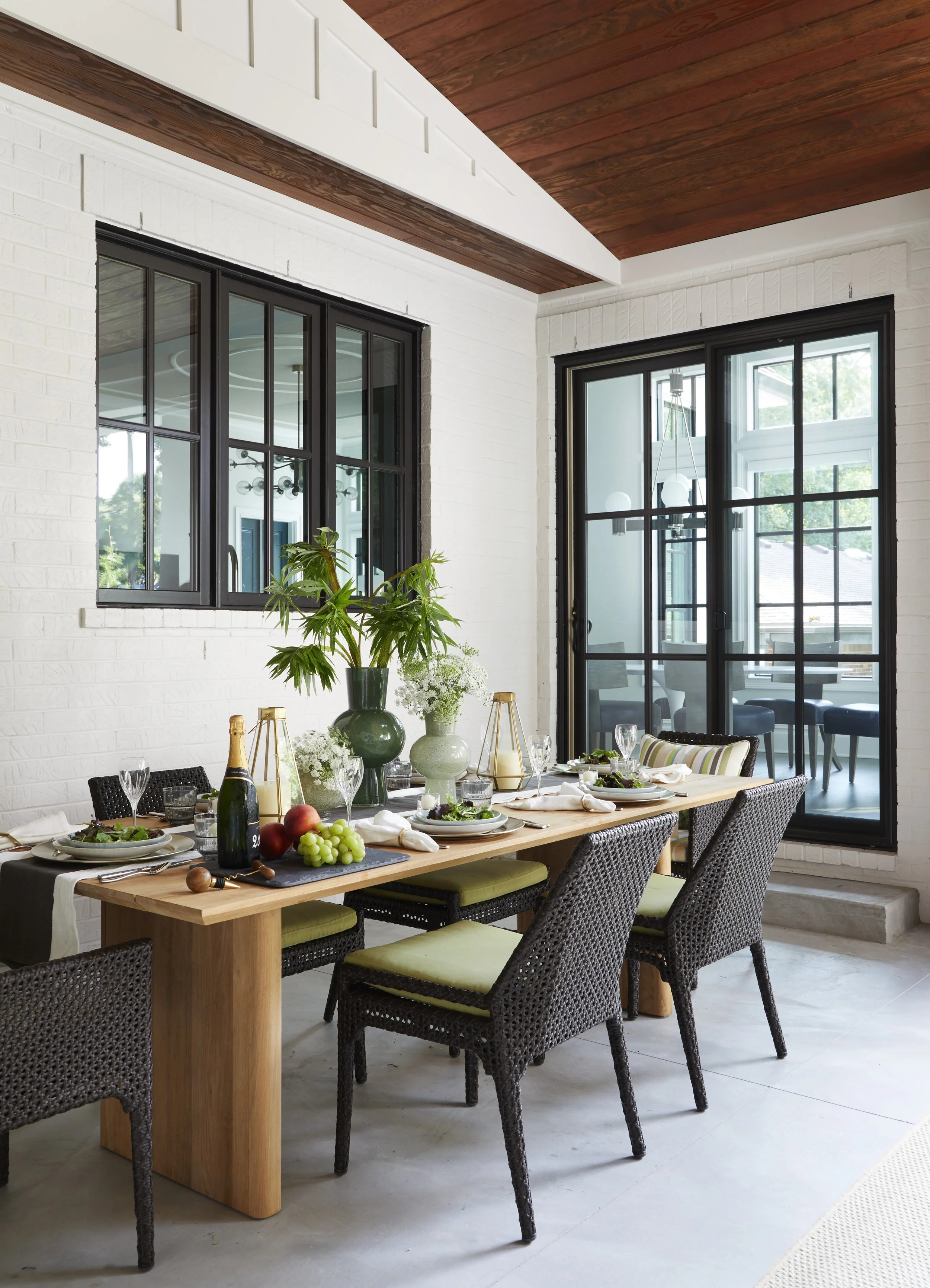A dining room with a wooden table set for a meal, surrounded by wicker chairs with green cushions. The table is decorated with wine bottles, drinks, fruits, bread, and a centerpiece of green and white vases with plants. Large windows and a sliding gl