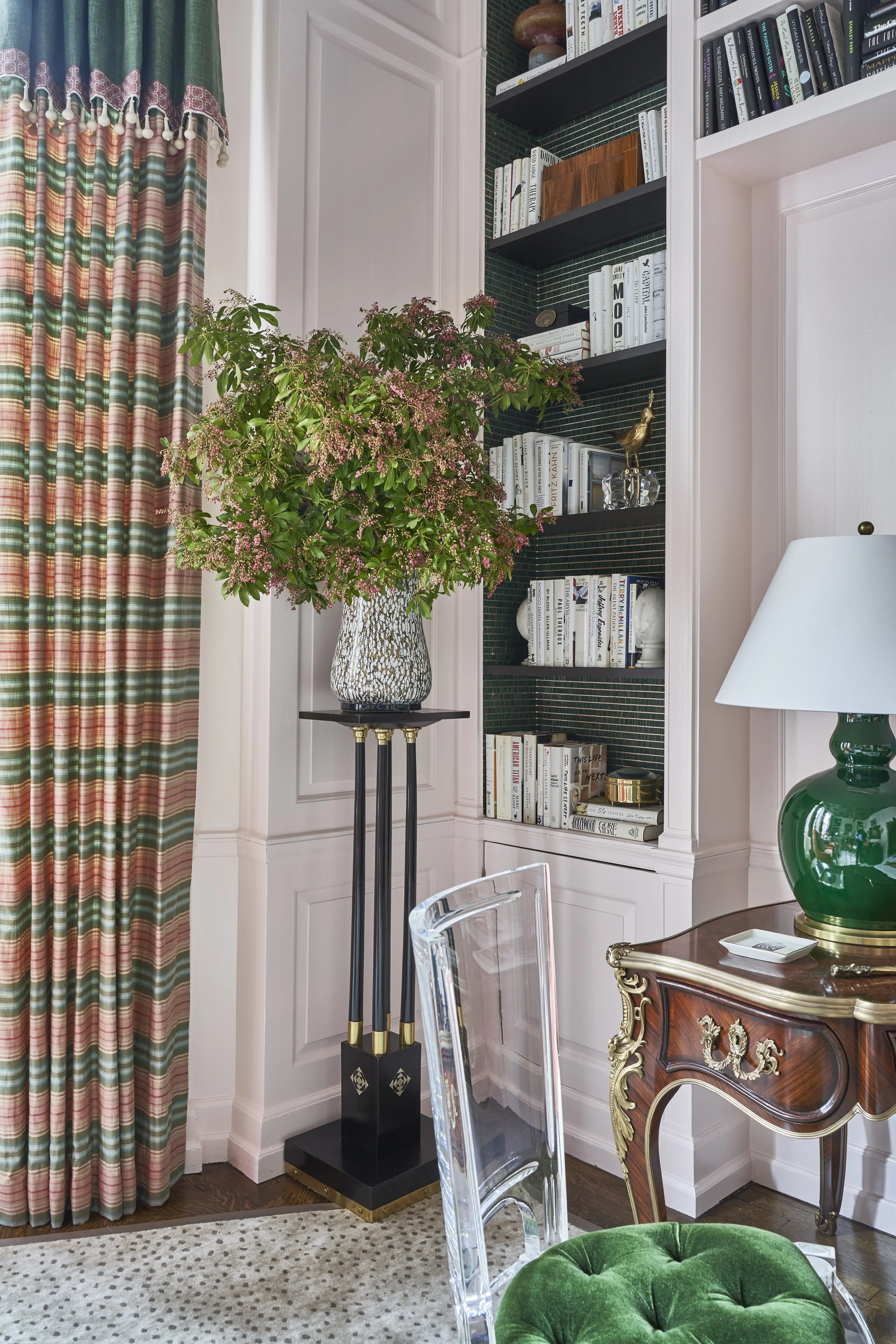 Interior view of a room with a colorful curtain on the left, a green leafy plant in a patterned vase on a black stand, a built-in bookshelf with various books, a decorative golden bird figurine, and a lamp on an ornate wooden side table. Part of a gl