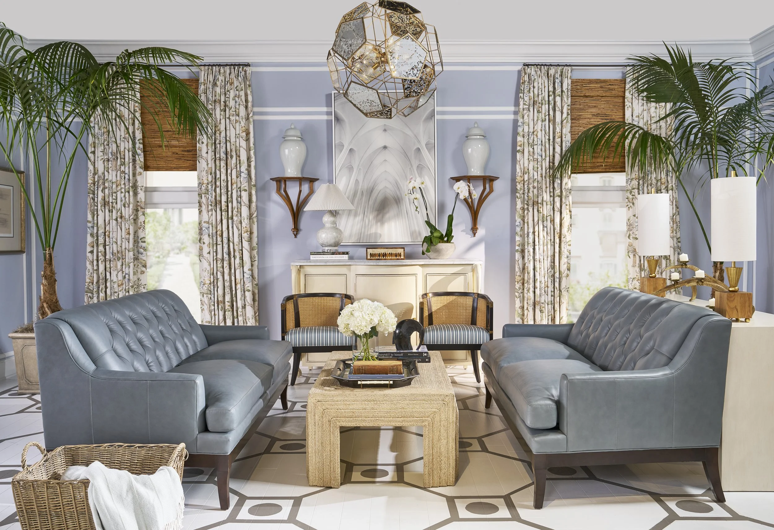 Living room with two gray leather sofas facing each other, a wooden coffee table in between, a white fireplace with decorative items, lush green plants on either side, curtains with a floral pattern, and a geometric hanging light fixture.