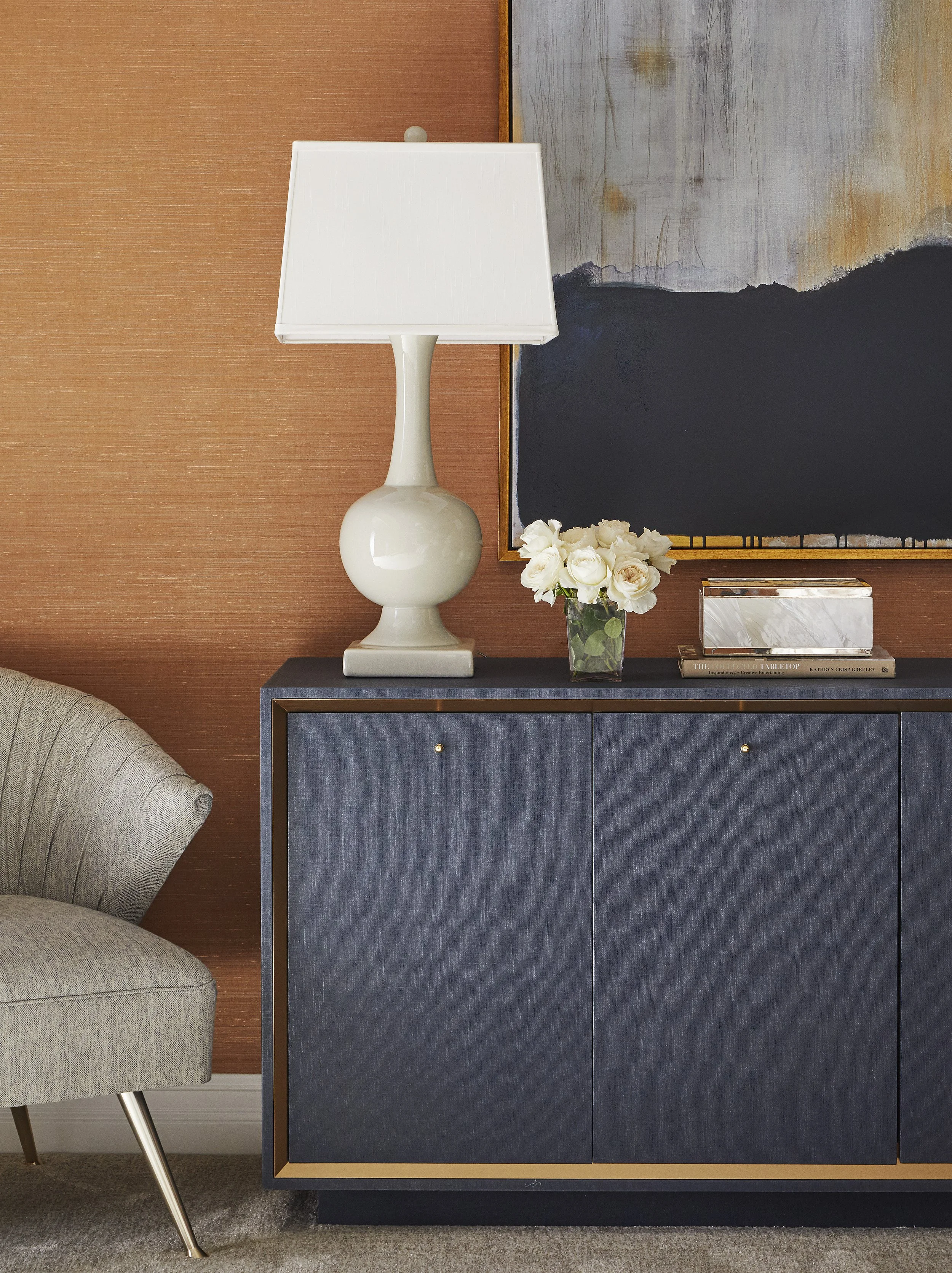Interior scene featuring a white table lamp with a rounded base and rectangular shade, a vase with white roses, and a dark blue cabinet with gold trim, against a two-tone wall with brown textured wallpaper and an abstract painting.